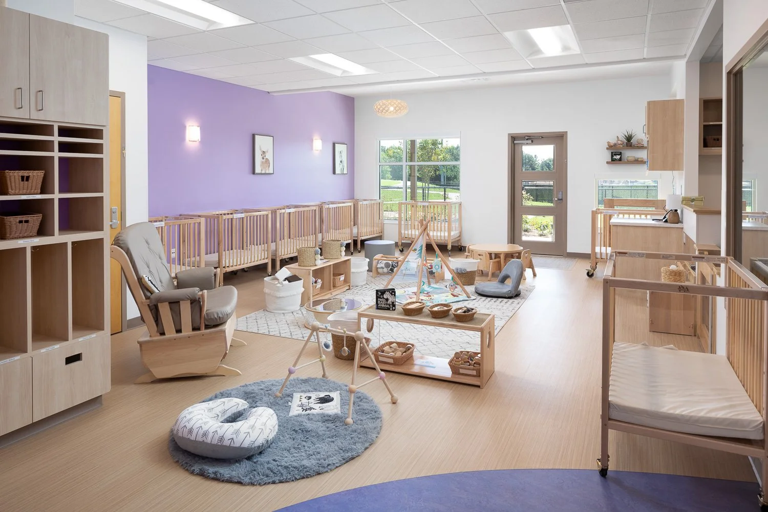 Infant classroom with cribs, soft play areas, low shelving, and a purple accent wall.