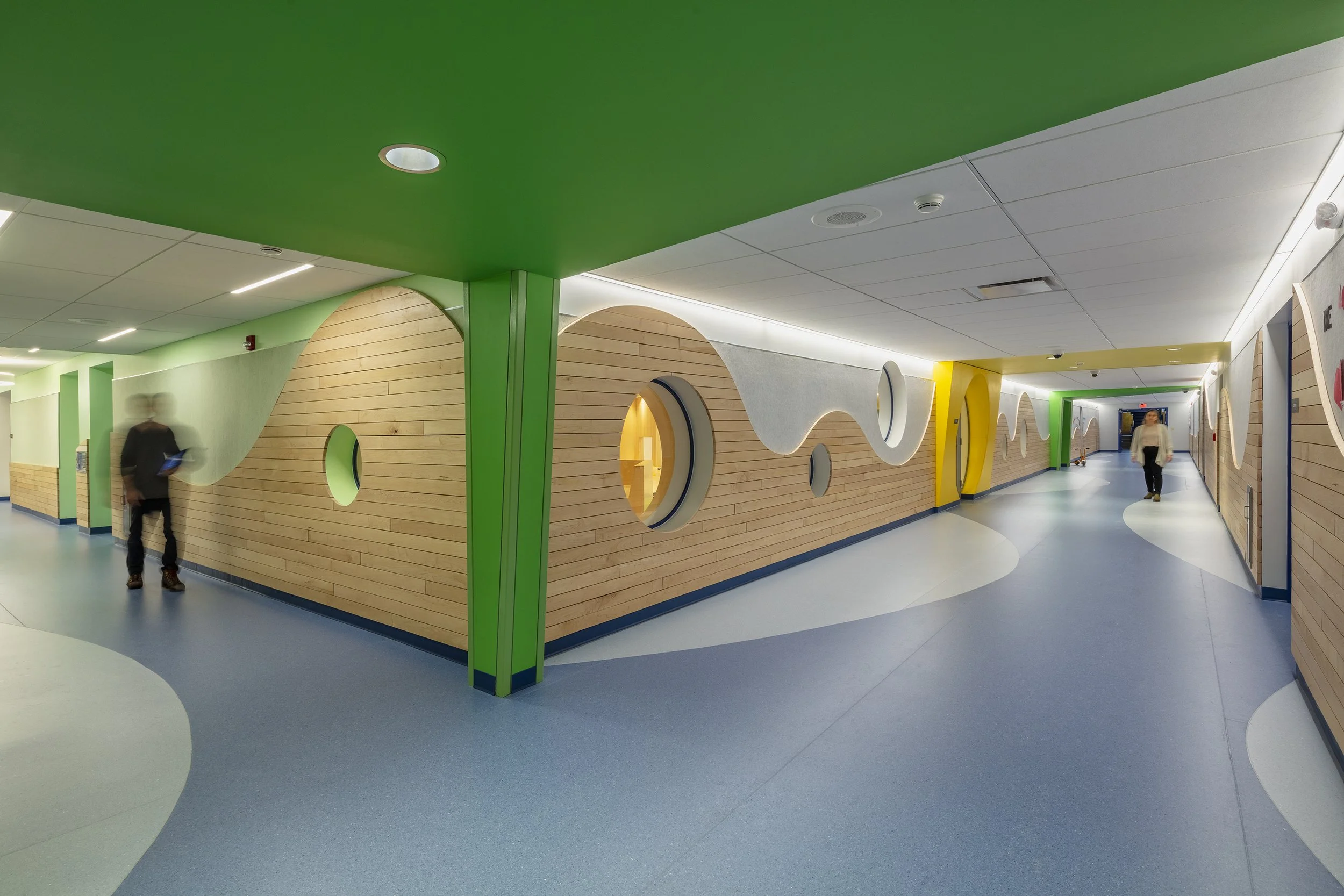 School corridor featuring curved blue flooring, wood-paneled walls with circular openings, and vibrant green and yellow ceiling accents.