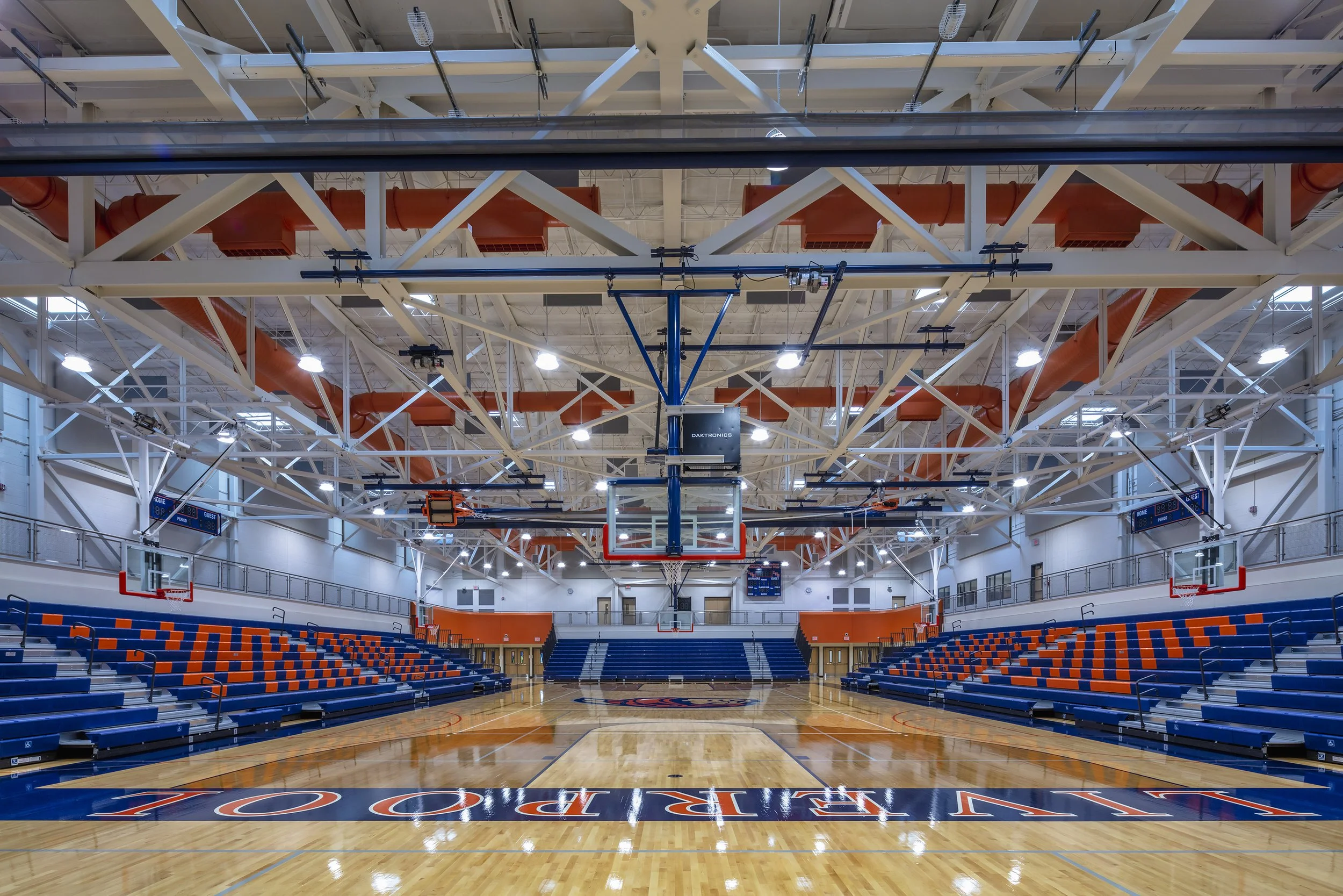 Liverpool High School gymnasium with basketball court, blue and orange bleachers, multiple hoops, and exposed roof structure.