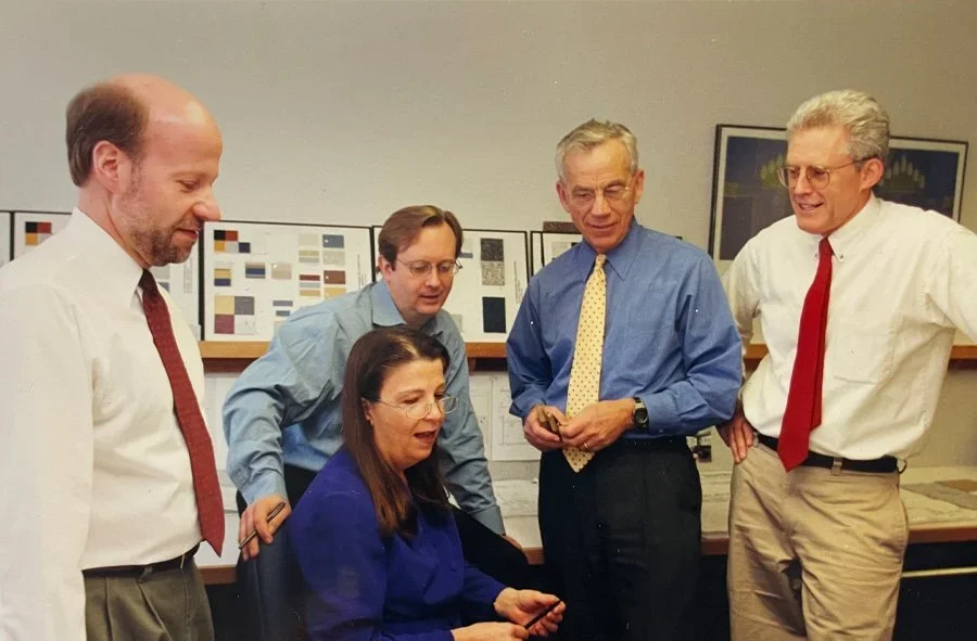 Five adults, four men and one woman, gather around a desk, looking at a smartphone. The woman is seated, wearing glasses and a blue blouse. The men are standing, wearing business casual attire with shirts, ties, and pants. The background features cha