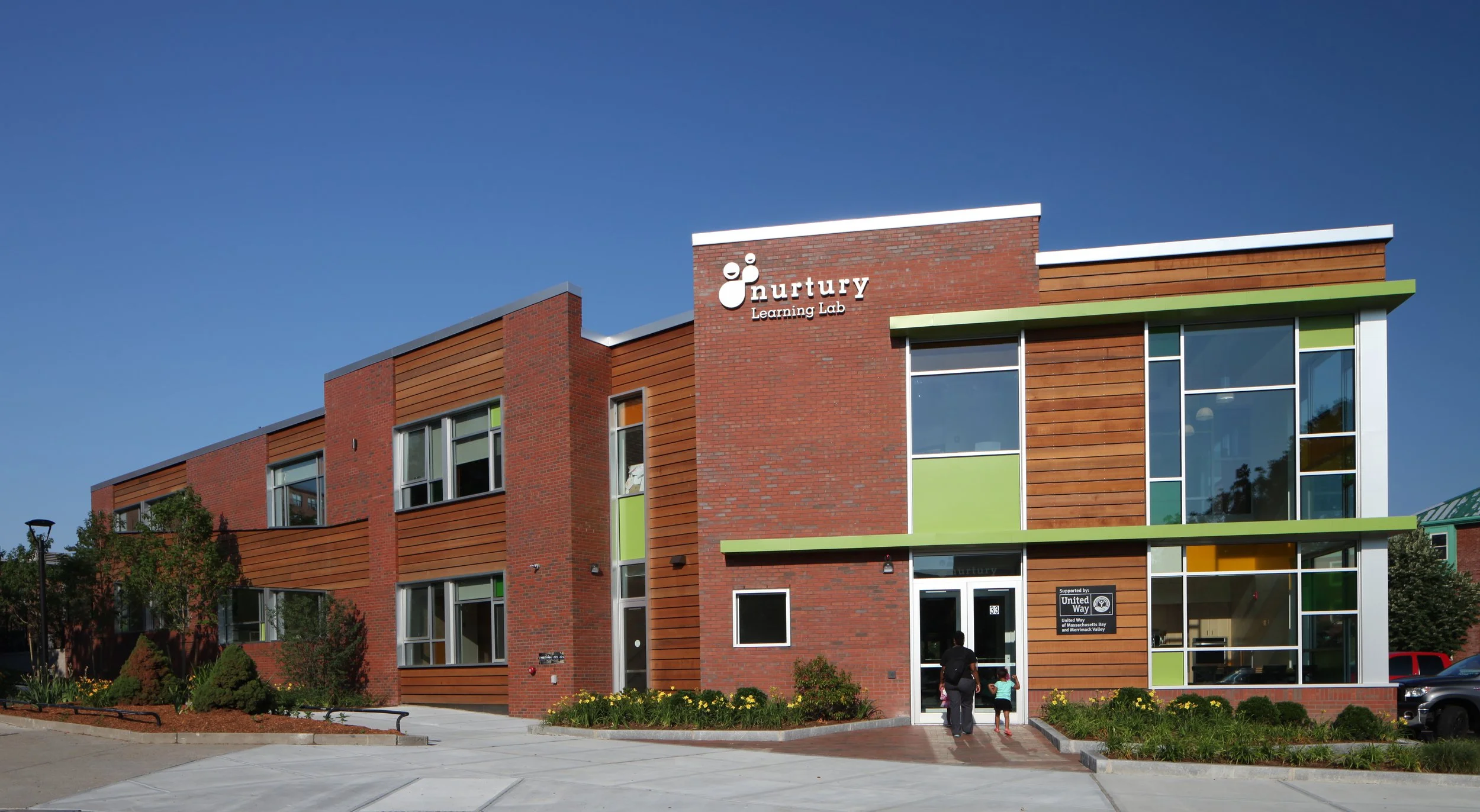 Exterior of Nurtury Learning Lab in Jamaica Plain, Boston, featuring brick, wood cladding, colorful window panels, and a landscaped entrance.