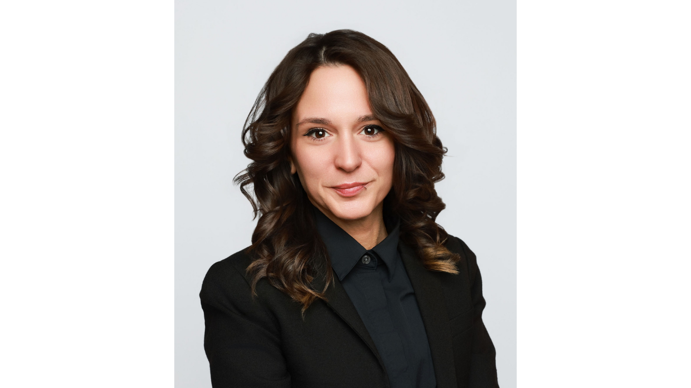 Professional headshot of a woman with dark wavy hair, wearing a black blazer and black collared shirt, photographed against a light gray studio background.