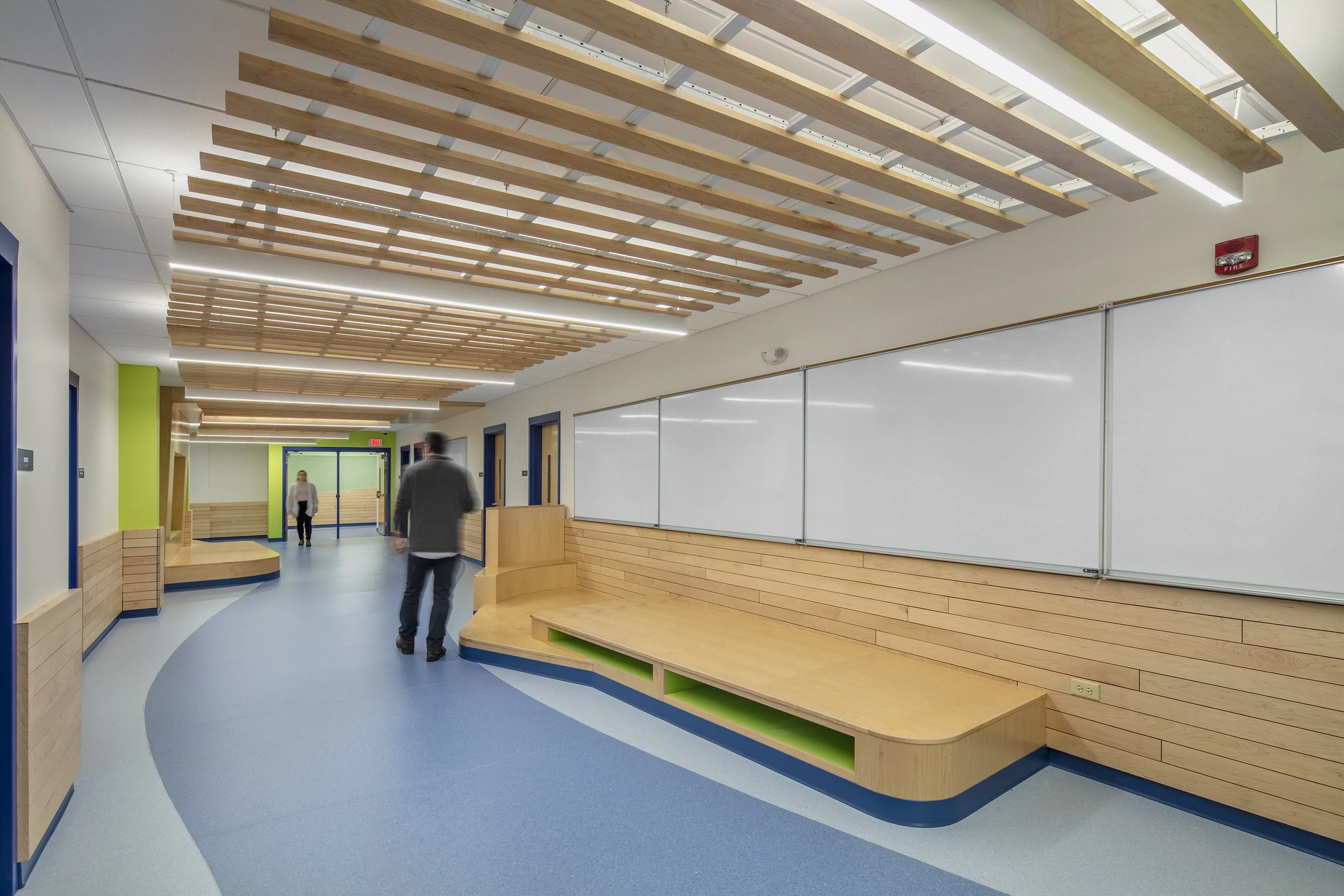 School corridor featuring built-in wood seating, wall-mounted whiteboards, suspended wood ceiling slats, and curved blue flooring.