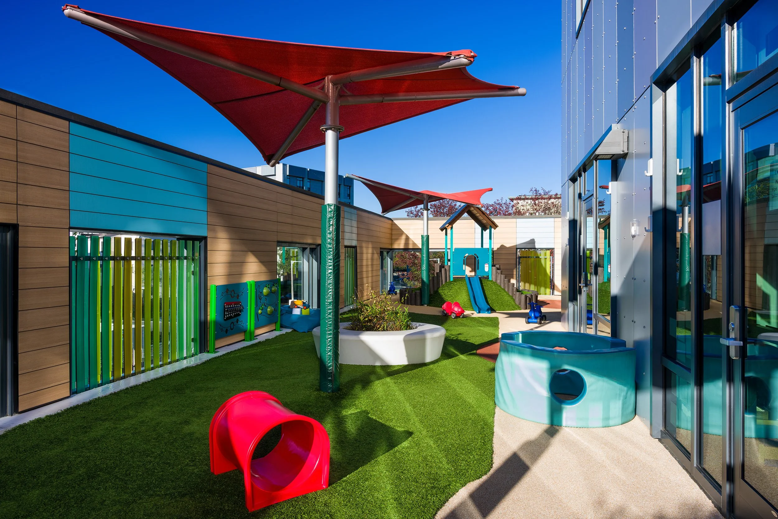 Outdoor early learning play area with artificial turf, red shade canopies, sensory play panels, and a small slide.