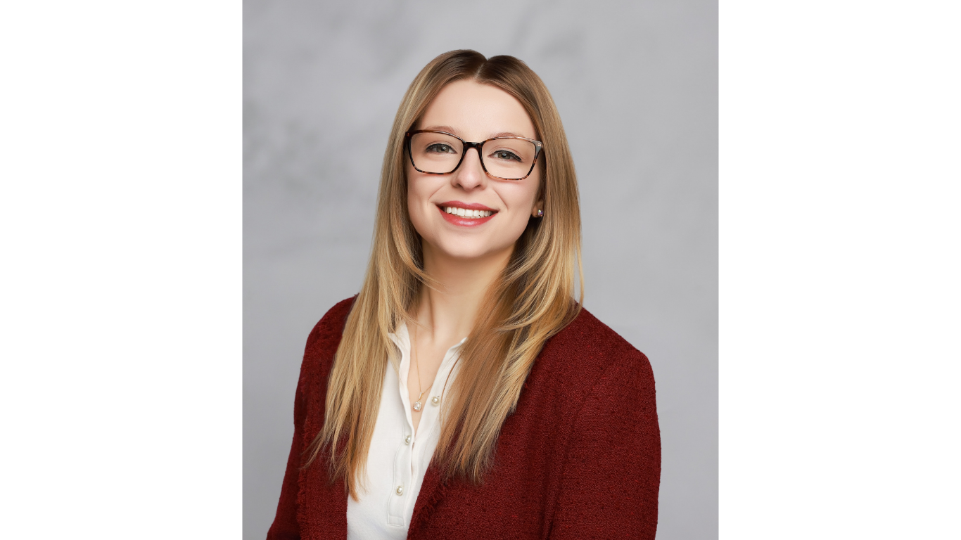 Professional headshot of a smiling woman with long blonde hair and glasses, wearing a white blouse and burgundy cardigan, photographed against a light gray studio background.