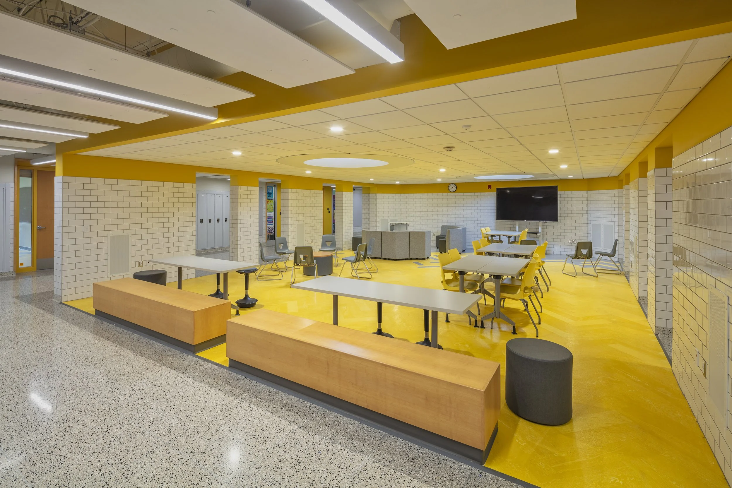 Student collaboration area featuring yellow flooring, white subway tile walls, wood benches, flexible seating, and suspended acoustic ceiling panels.