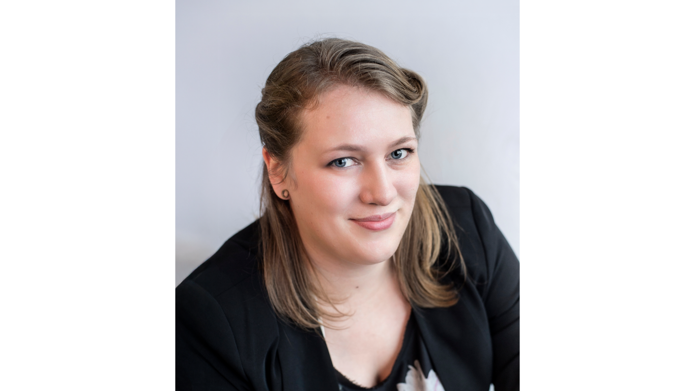 Professional headshot of a woman with light brown hair, wearing a black blazer over a patterned top, smiling at the camera against a light gray studio background.
