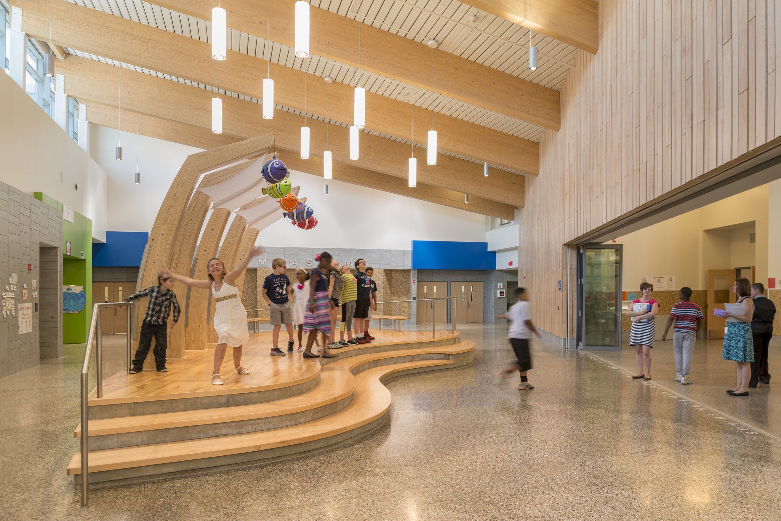 Central school gathering space with tiered seating, exposed wood structure, and students using the space for activities.