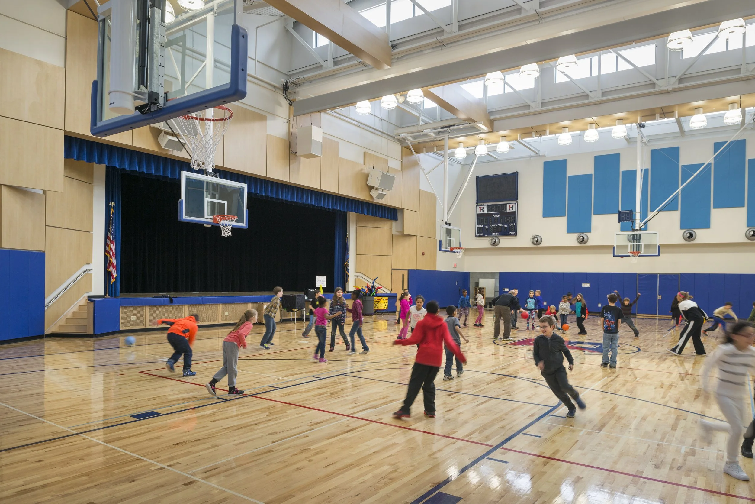 Elementary school gymnasium with a stage, basketball courts, abundant daylight, and students participating in physical activities.