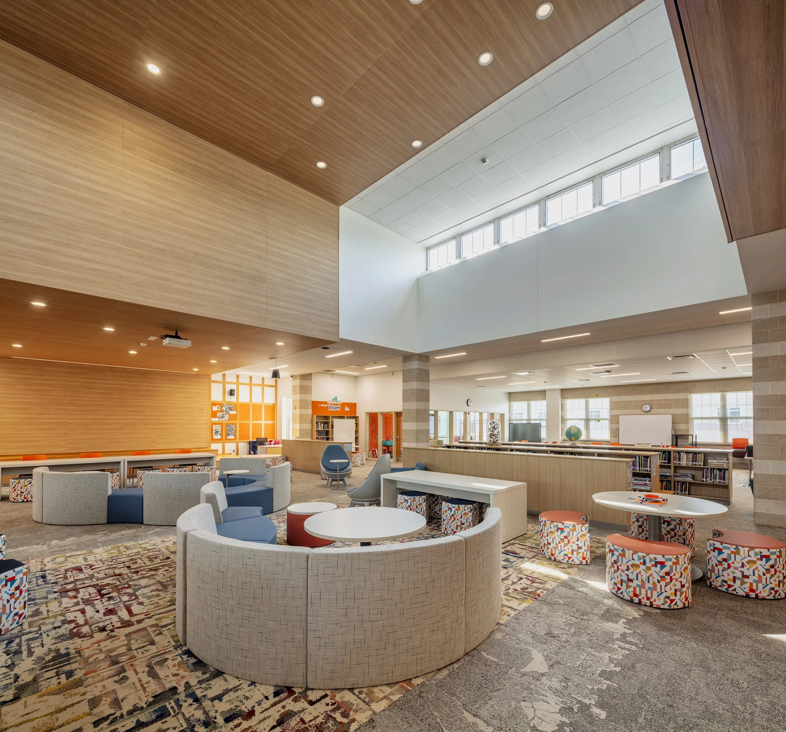 Double-height library media center with clerestory windows, wood ceiling panels, curved seating, and colorful furniture for individual and group learning.