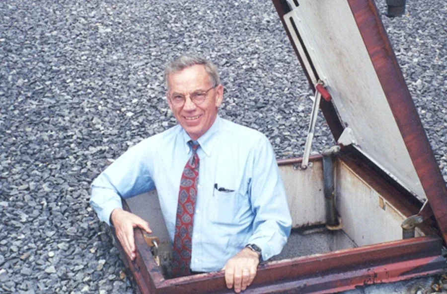 An elderly man in glasses, a light blue shirt, and a patterned tie standing inside an open industrial hatch that is embedded in a gravel surface.