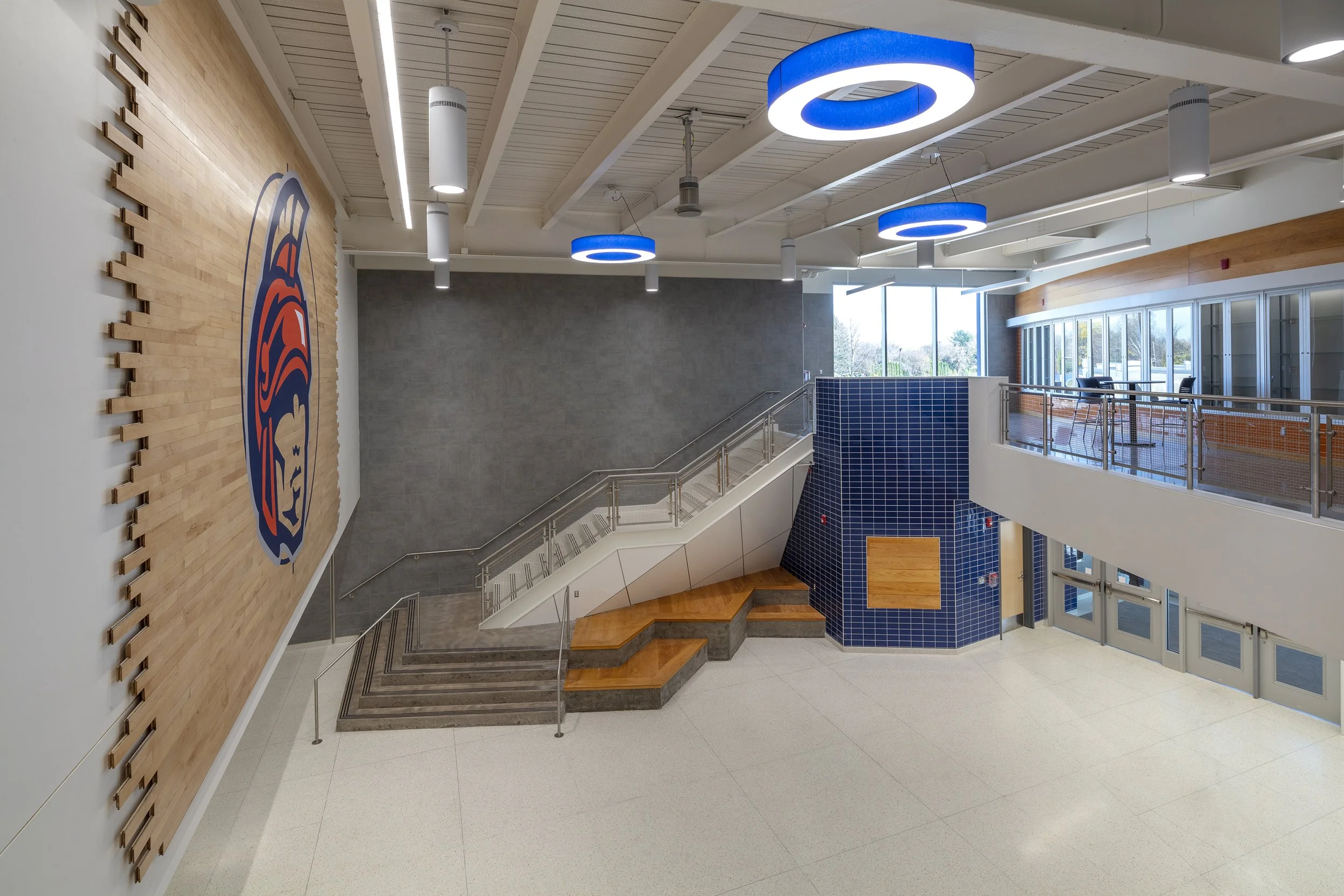 Upper level view of the athletics lobby featuring Warriors branding, tiered seating, staircase, and blue circular lighting.
