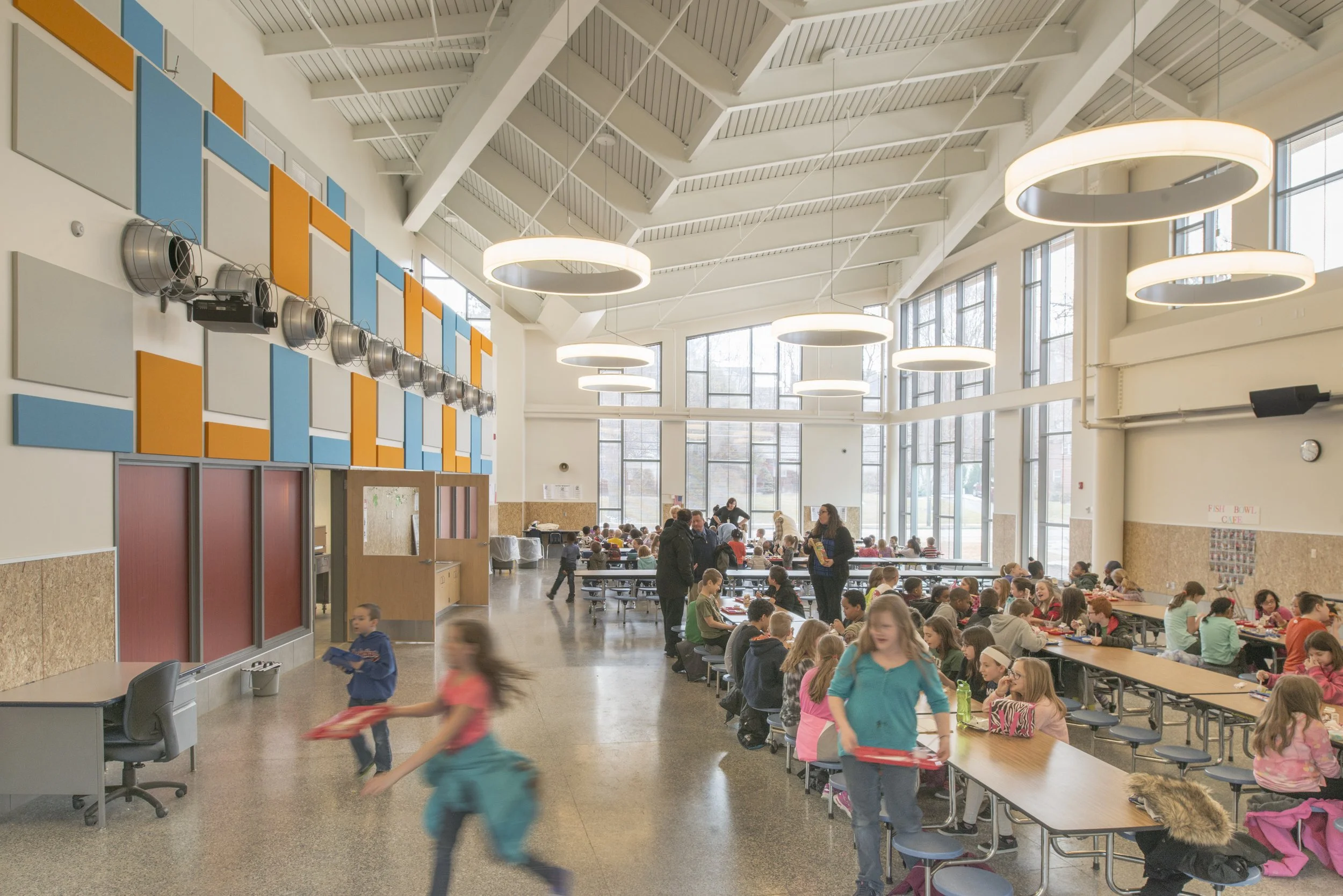 Bright elementary school cafeteria with large windows, colorful acoustic panels, and students gathering for lunch.