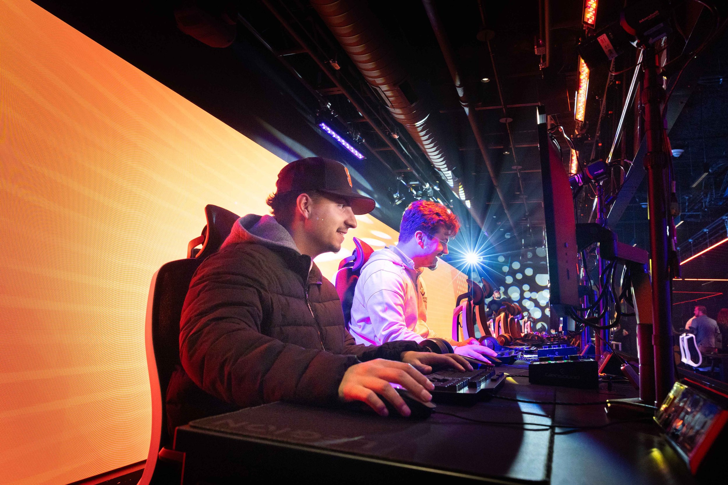 Gamers play computer games during the grand opening of a gaming and esports center, seated at gaming stations beneath colorful lighting and a large digital display.