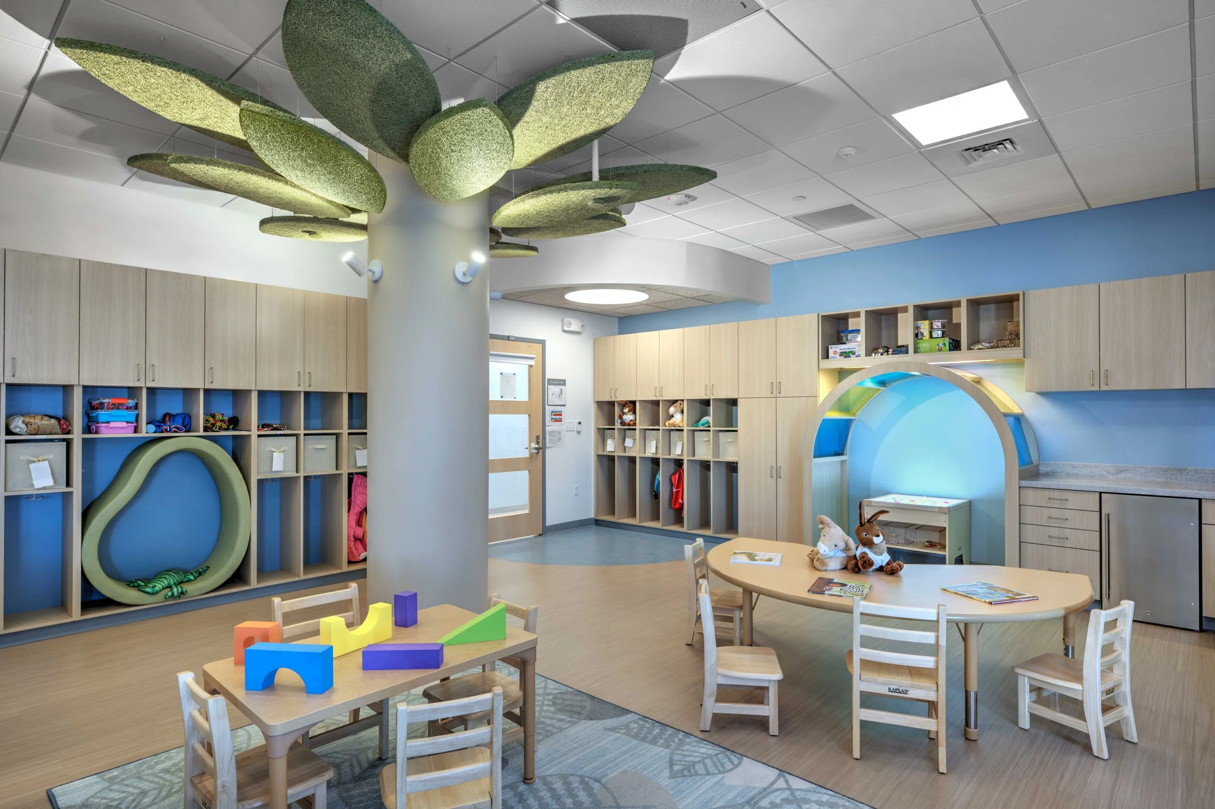 Early childhood classroom featuring a tree-inspired acoustic ceiling element, natural wood finishes, built-in storage, and a semi-enclosed reading nook designed to support exploration and self-regulation.