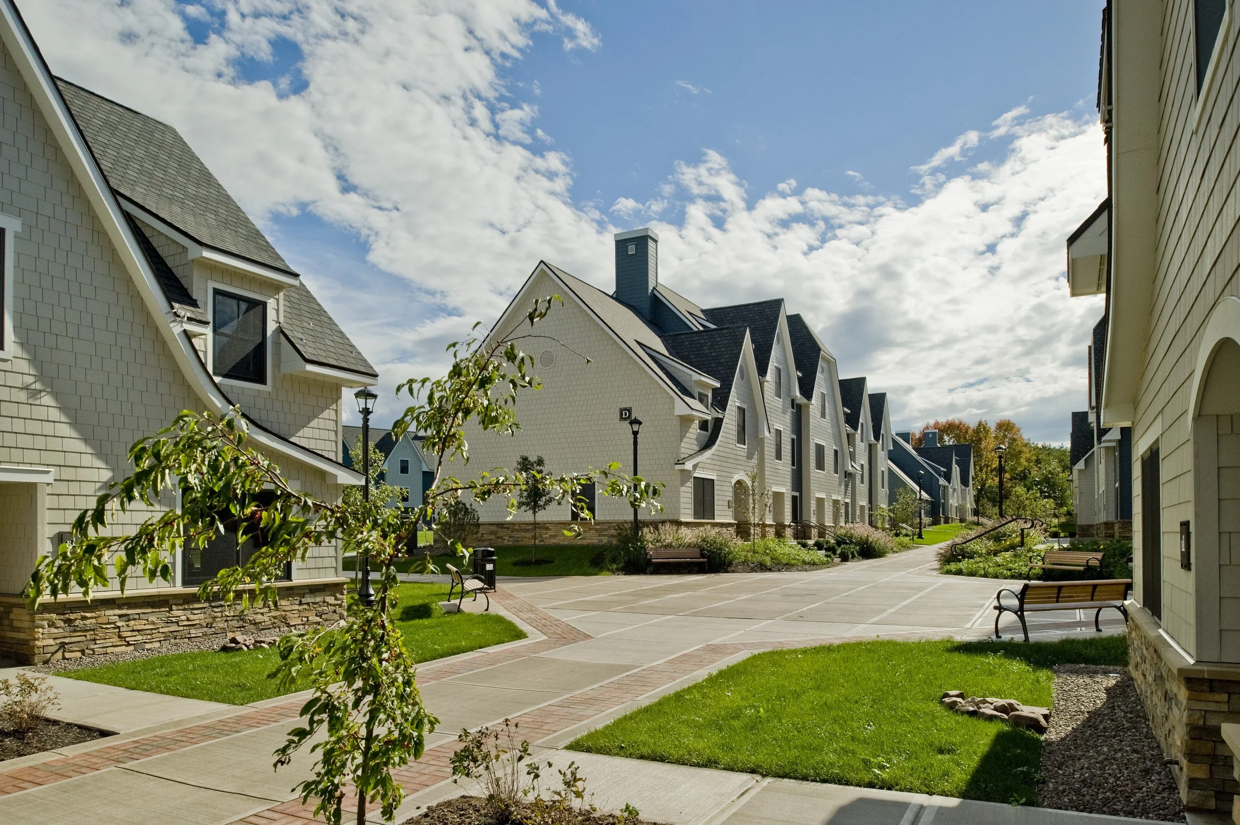 Pedestrian walkway through a residential apartment community featuring landscaped courtyards, benches, and multi-family buildings.