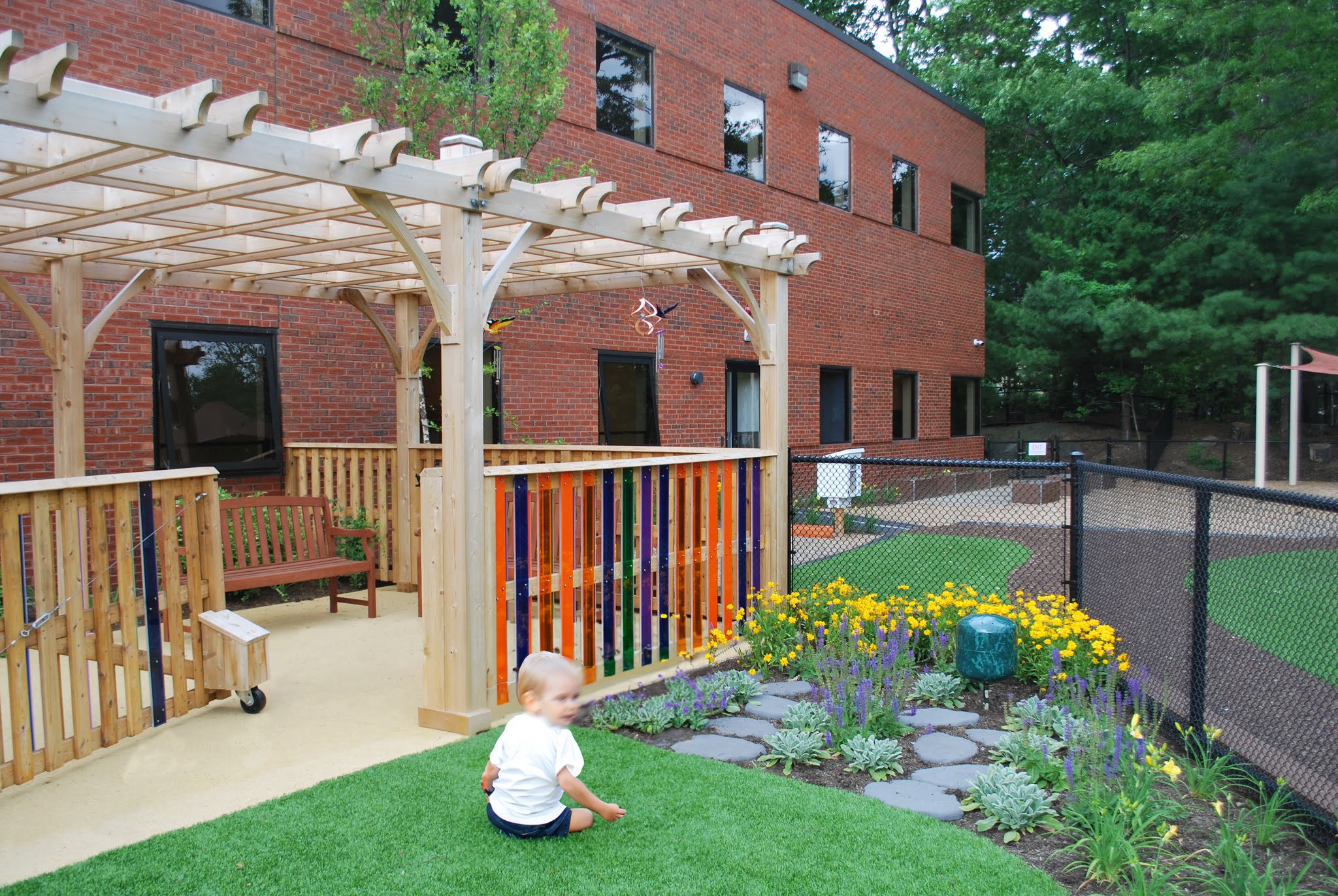 Childcare outdoor space with a wood pergola, colorful fencing, sensory garden, and lawn play area.