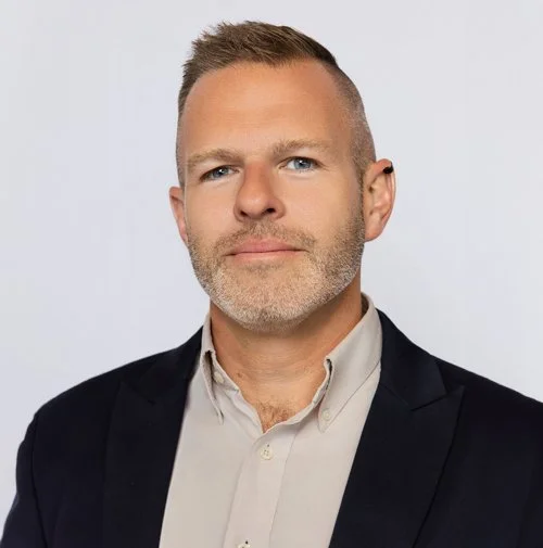 A person with short light brown hair and a closely trimmed beard wears a light button-down shirt beneath a dark blazer, looking toward the camera against a light gray background.