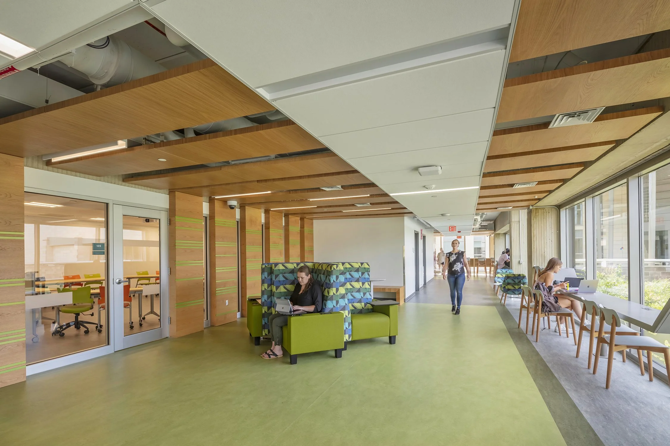 Bright student collaboration area with flexible seating, study tables, wood ceiling elements, and large windows overlooking the courtyard.