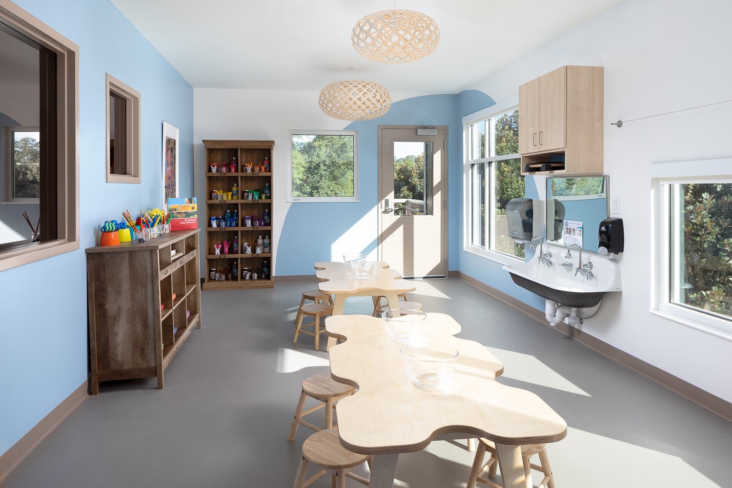 Bright art classroom with child-sized worktables, art supply storage, sinks, and large windows overlooking the landscape.