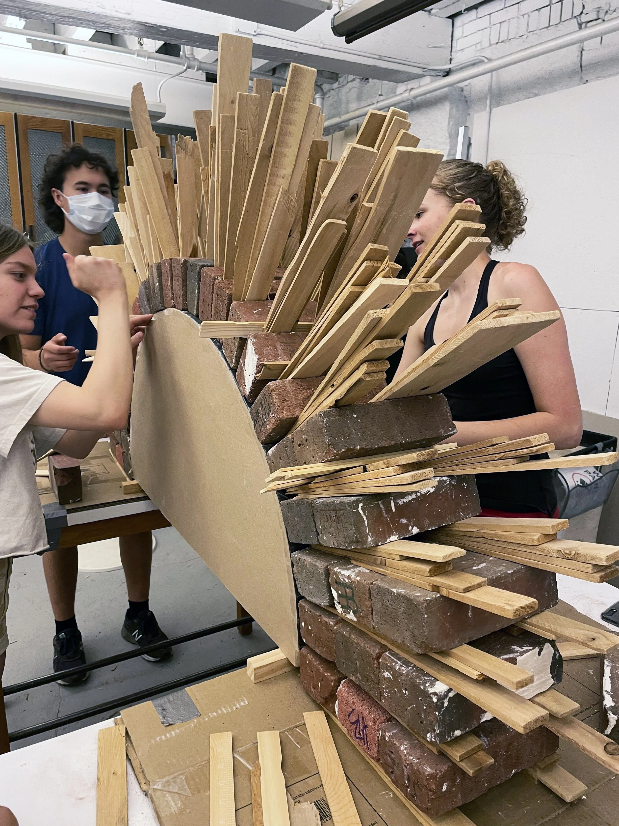 High school students collaborate on a hands-on masonry workshop, building a mock wall using reclaimed bricks and wood shims.