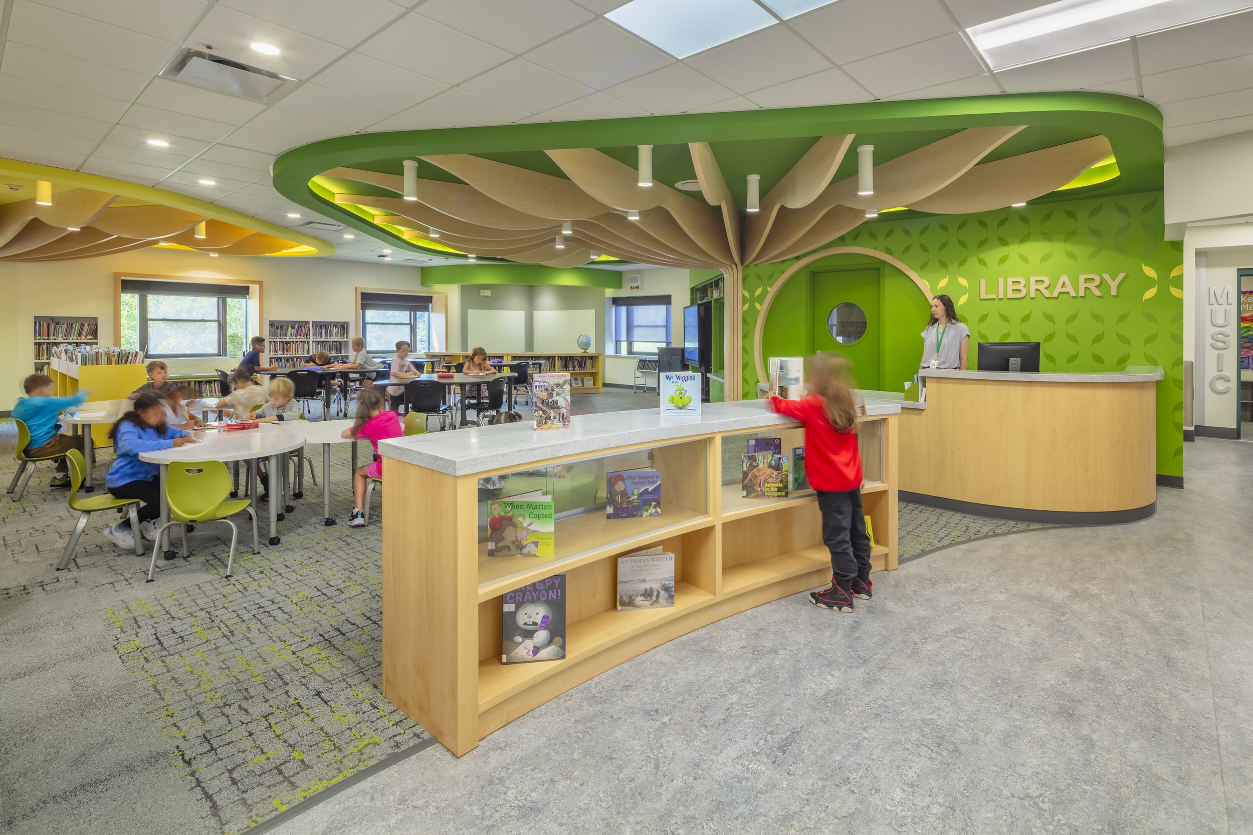 Elementary school library with students working at tables beneath colorful tree-inspired ceiling features.