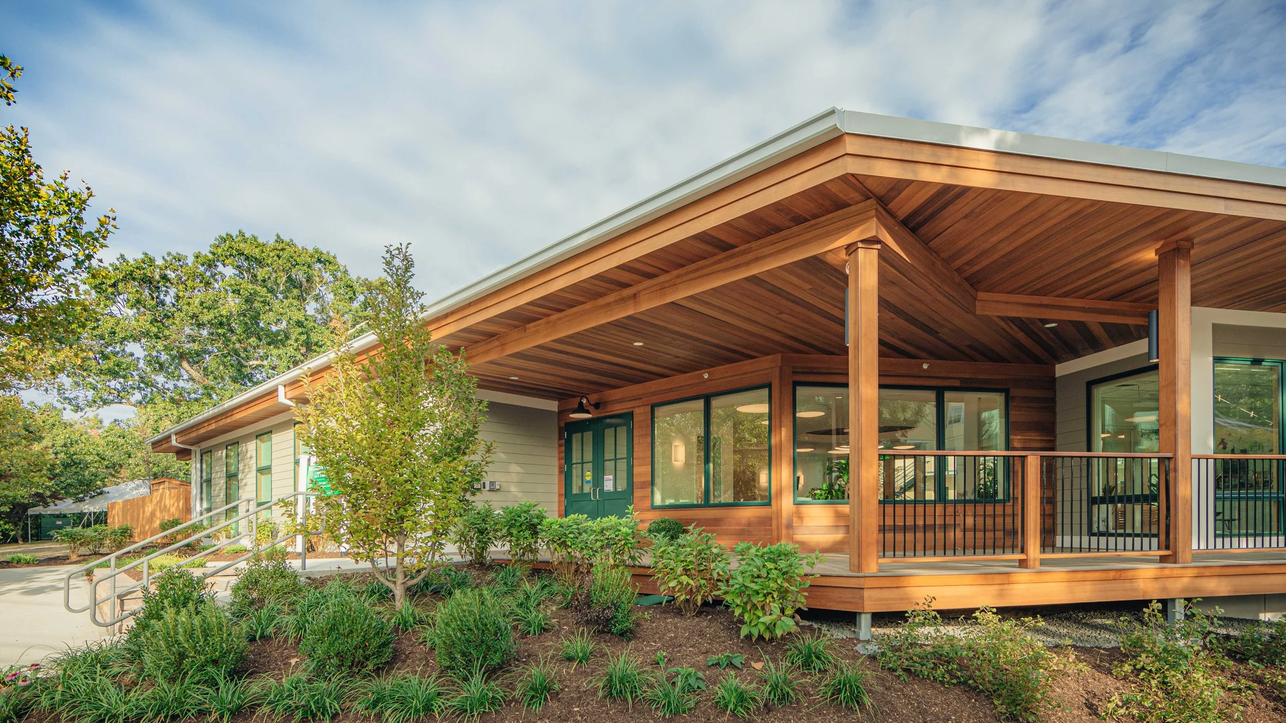 Covered entrance to a contemporary early childhood school featuring warm wood soffits, landscaped gardens, accessible walkways, and large windows into learning spaces.