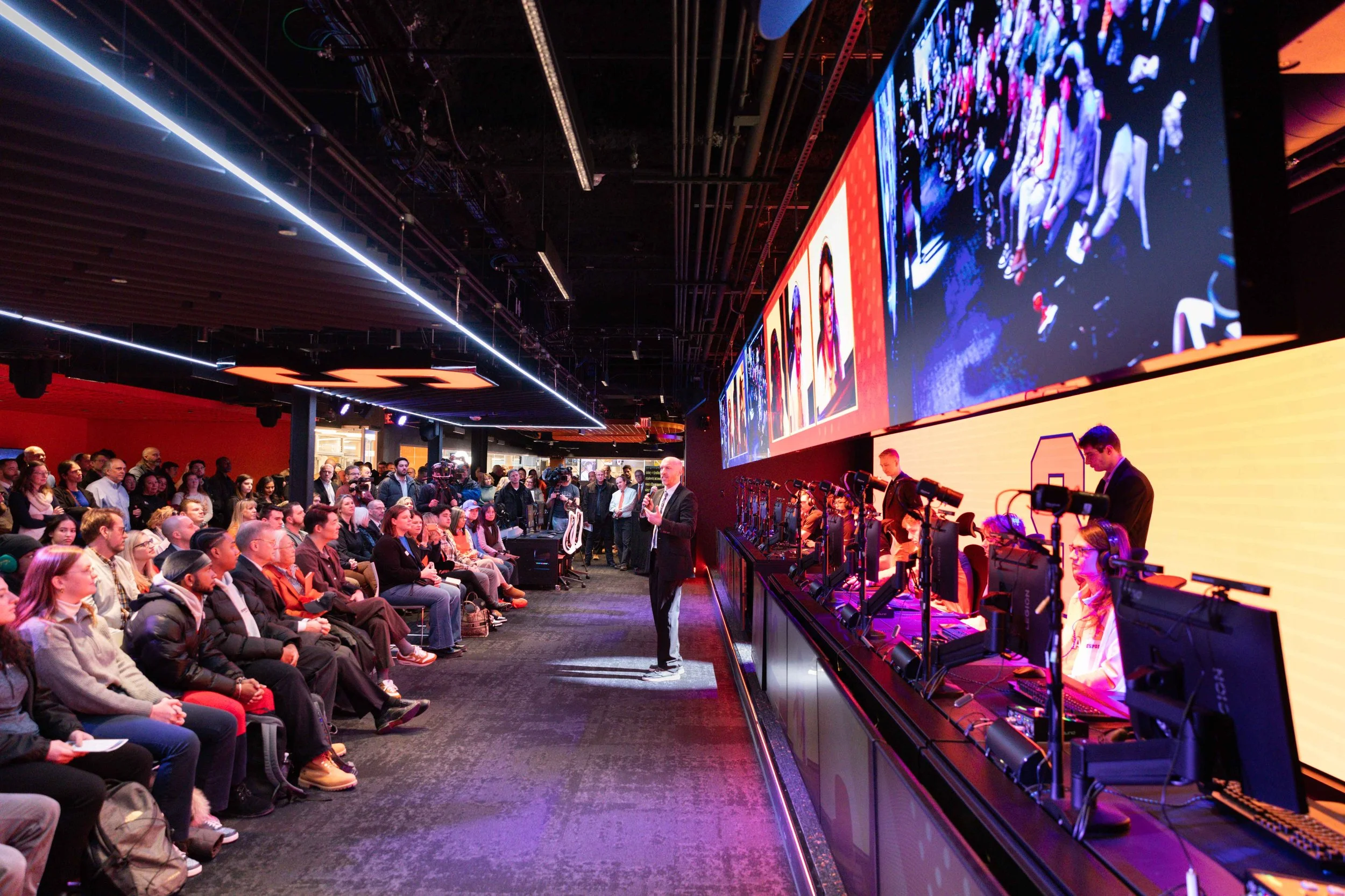 Guests gather for the grand opening of a gaming and esports center as a speaker addresses the audience beside gaming stations and large digital screens displaying live event content.
