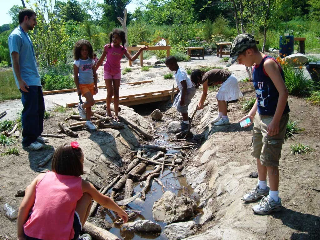 Children explore a natural dry stream bed with rocks, logs, and shallow water while an adult supervises at the Boston Nature Center's Nature Nook.