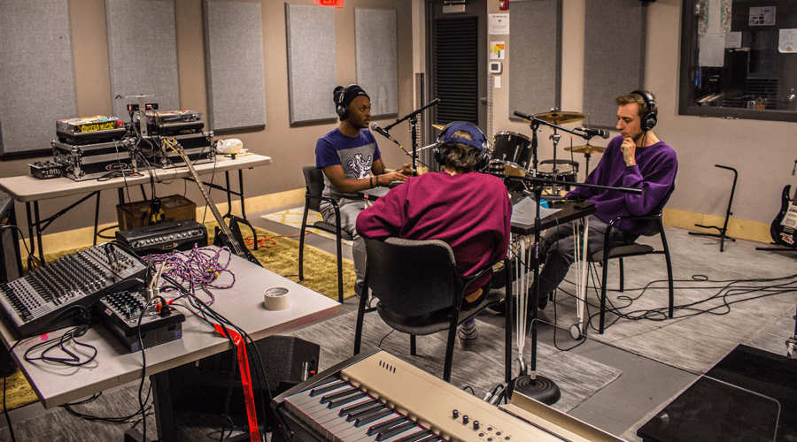 Teens work together in a recording and music production space with microphones, instruments, mixing equipment, and acoustic wall panels.