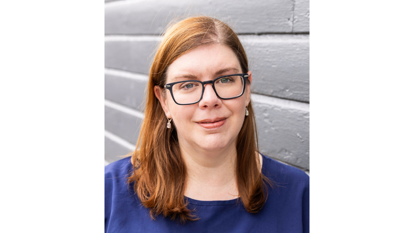 Professional headshot of a woman with red hair and glasses, wearing a blue top, standing in front of a gray wall and looking toward the camera.