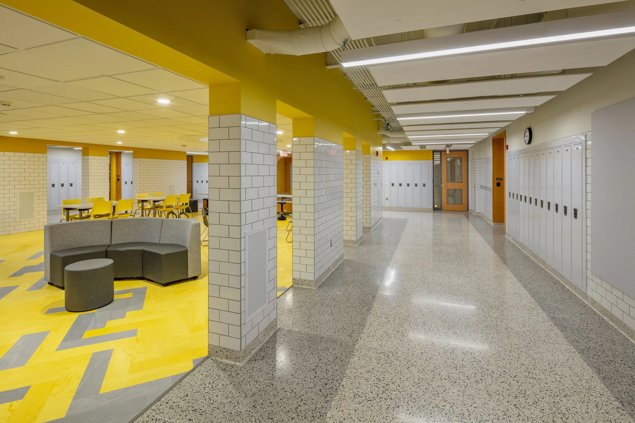 School corridor featuring white lockers, terrazzo flooring, and an adjacent student lounge with yellow flooring and gray upholstered seating.