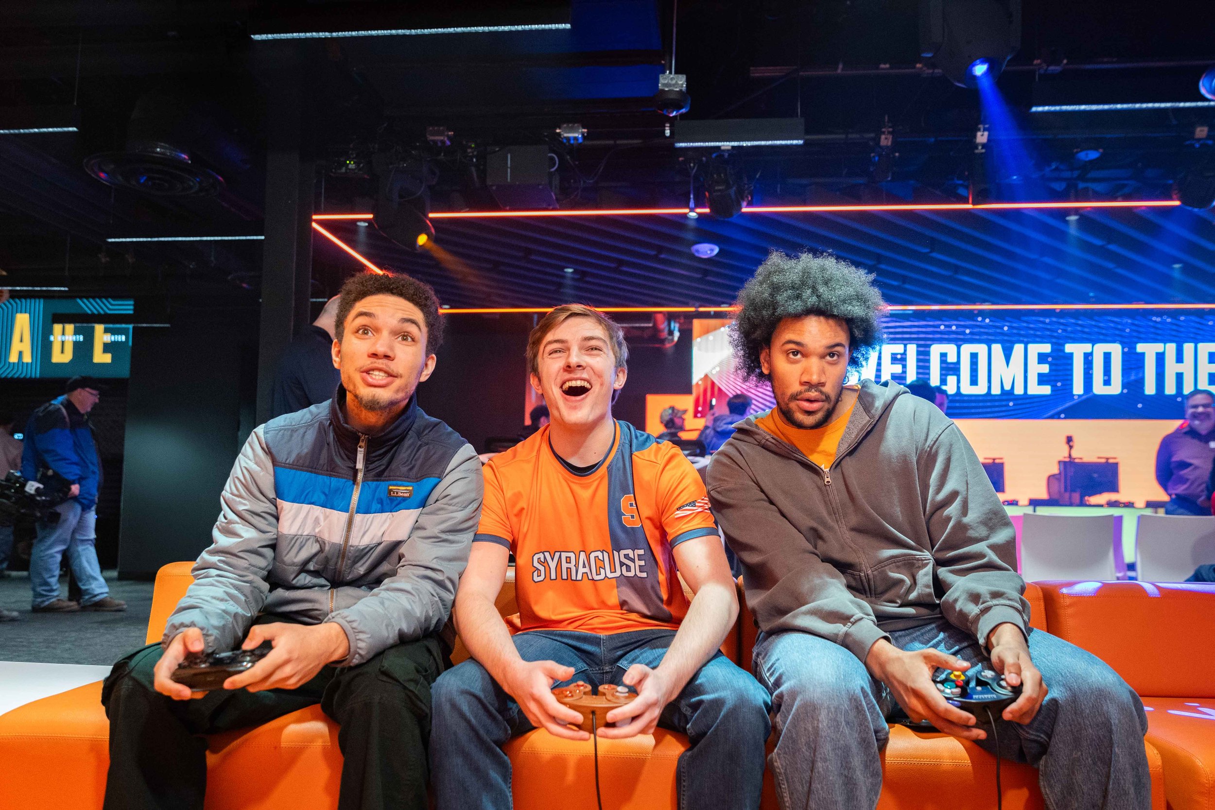 Three gamers play a video game with handheld controllers during the grand opening of a gaming and esports center, seated on a couch beneath colorful lighting and digital displays.