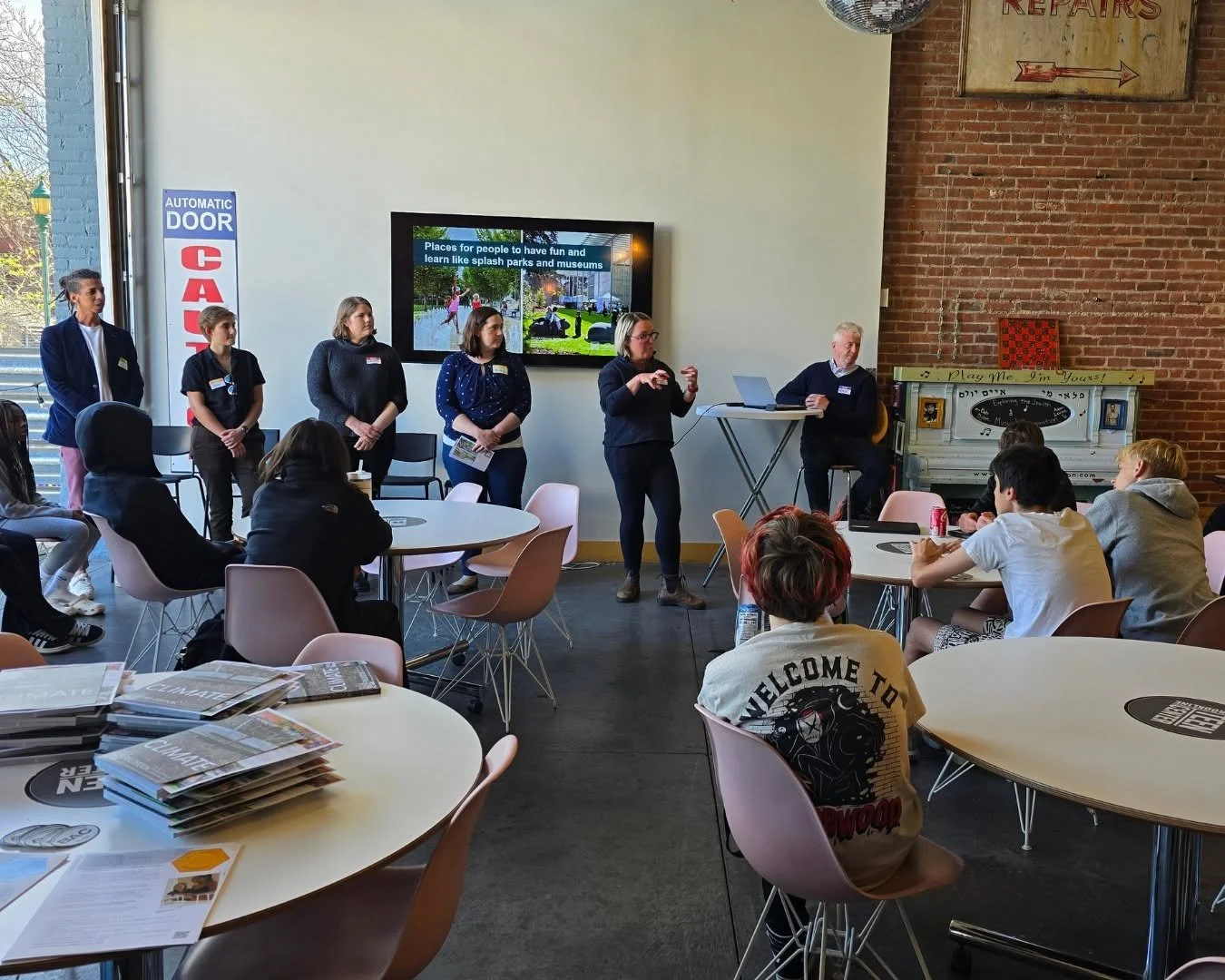 Students participate in a discussion at the Brookline Teen Center, seated at round tables in a flexible gathering space with exposed brick walls and an open garage door.