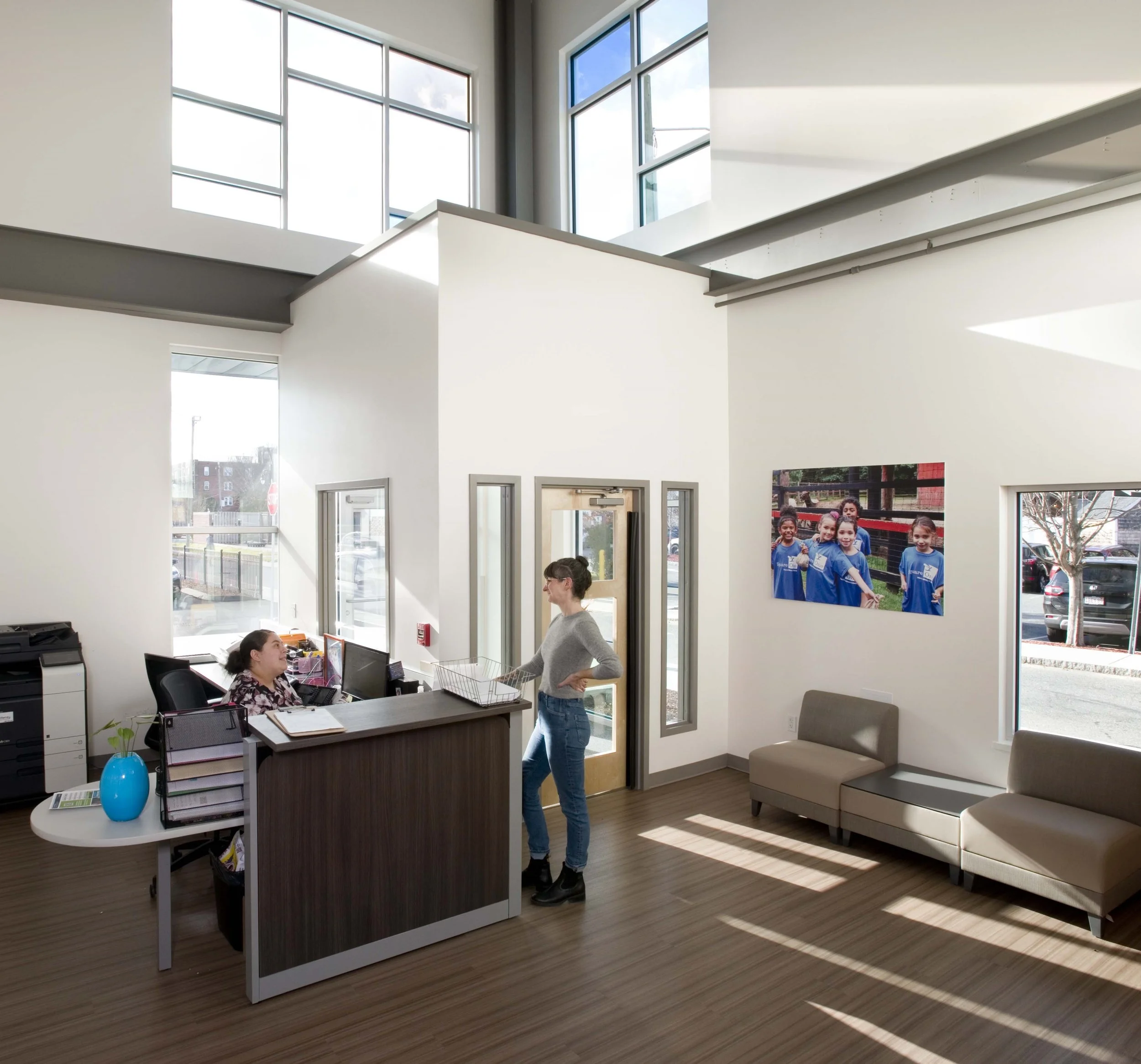 Sunlit reception and waiting area with high ceilings, large windows, and staff speaking at the front desk.
