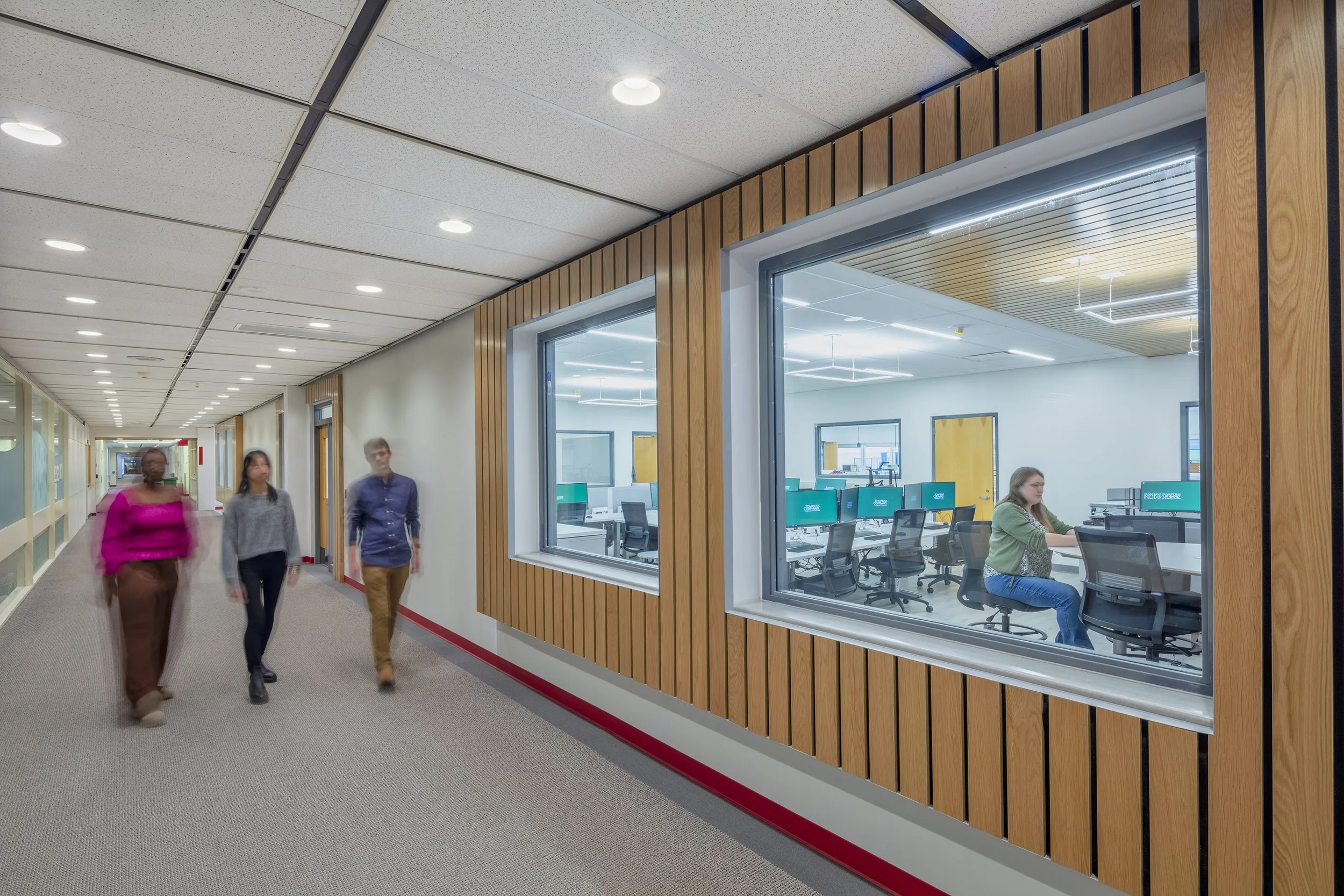 Academic corridor with interior windows providing visibility into adjacent learning spaces and wood wall detailing.
