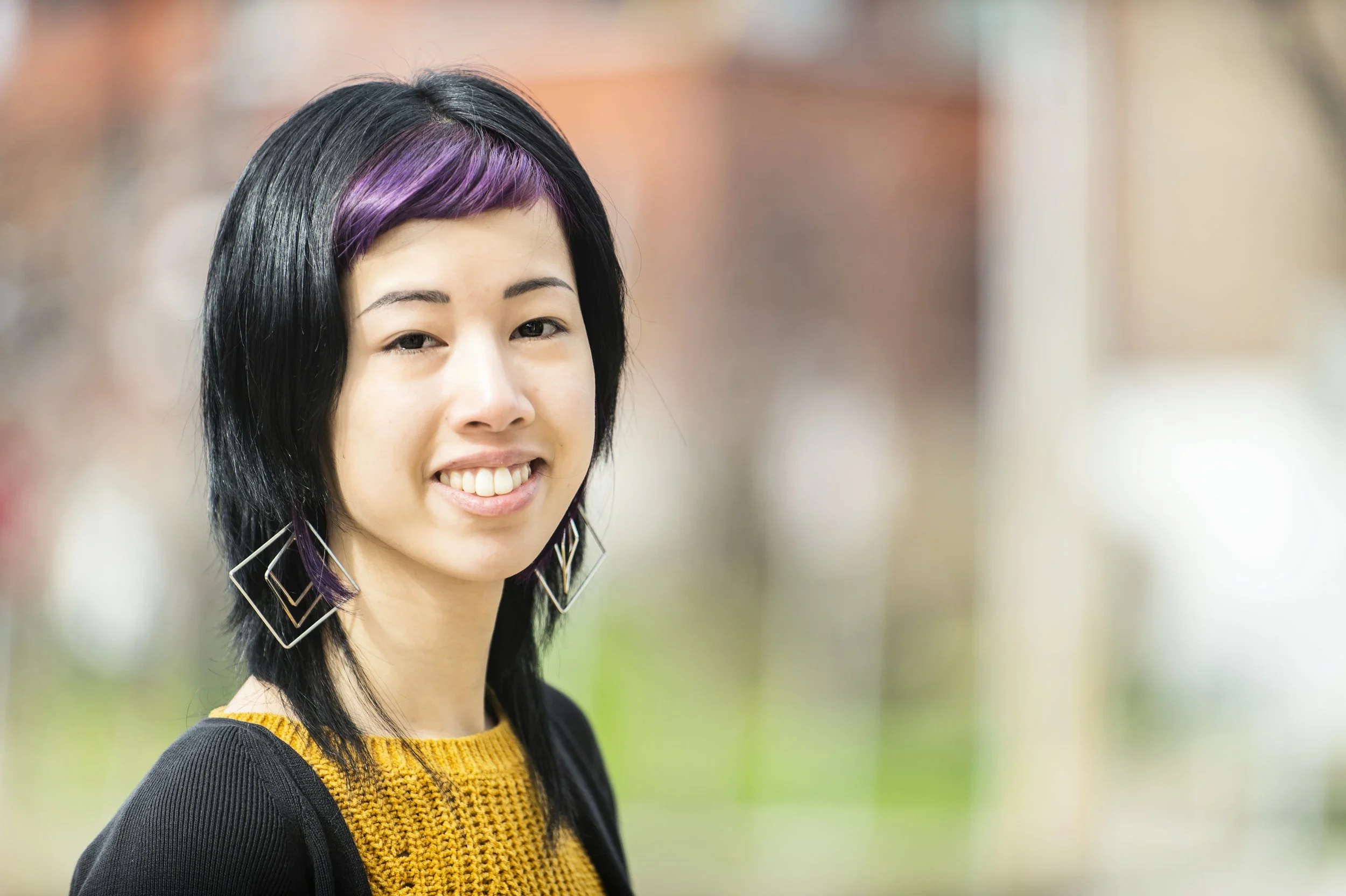 A person with black hair featuring purple highlights wears a mustard yellow knit top, black cardigan, and geometric earrings, smiling toward the camera against a softly blurred outdoor background.
