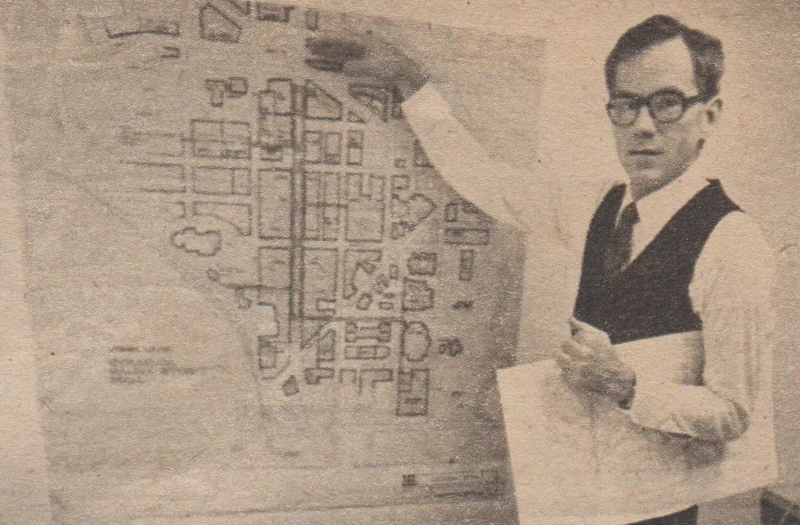 A man in glasses, a white shirt, and a vest points to a detailed map or blueprint on a wall with a pencil, while holding a paper in his other hand.