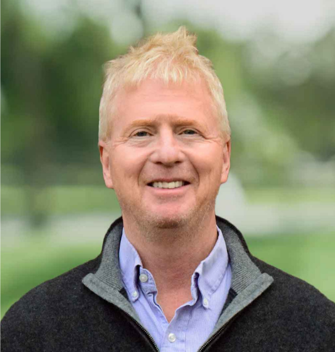 Portrait of a smiling man with blonde hair, wearing a blue button-up shirt and a dark zip-up jacket outdoors with a blurred green park background.