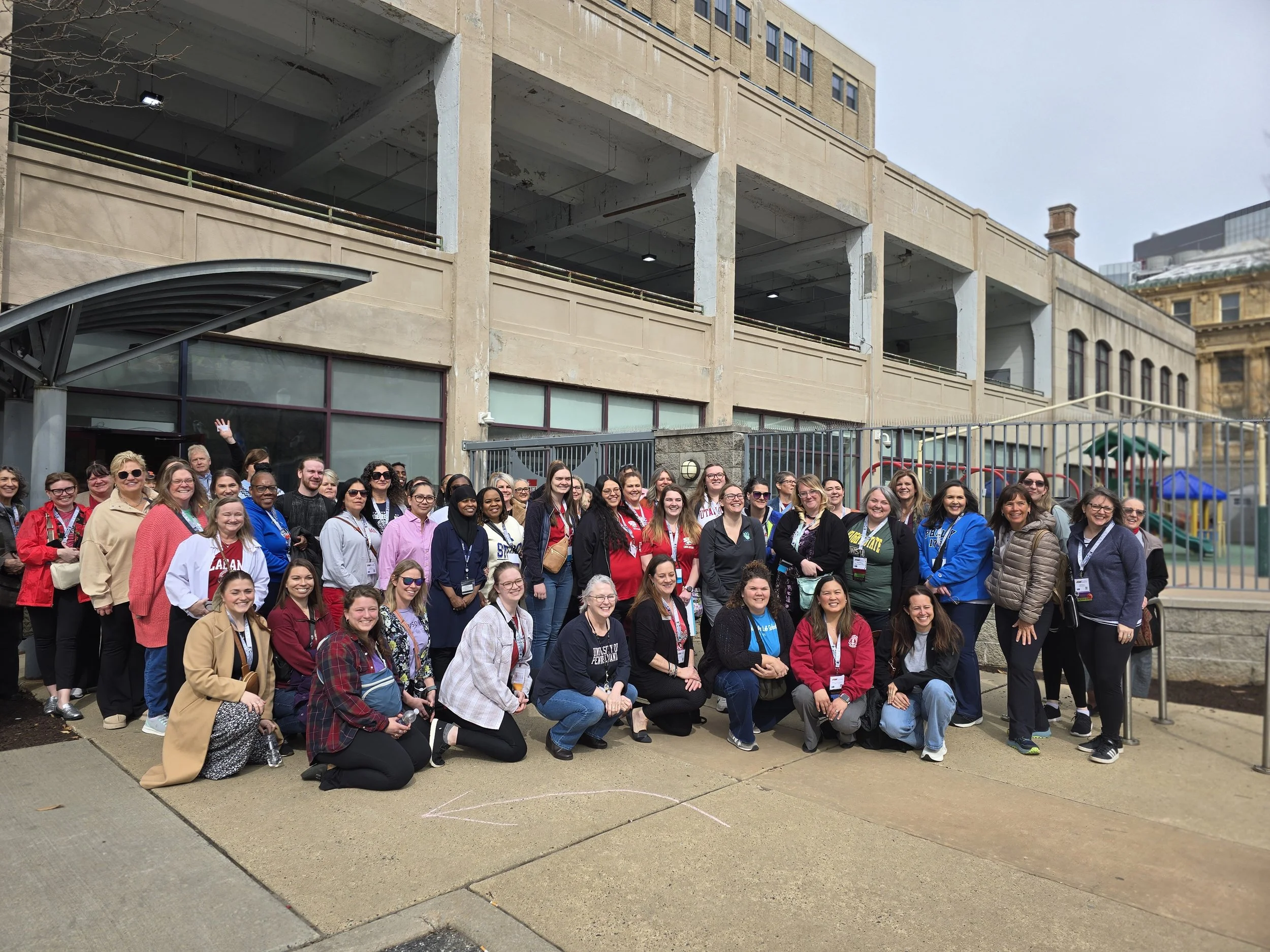 Group of conference attendees gathered outdoors for a site tour, posing together on a sidewalk beside a fenced playground and an urban child care facility.