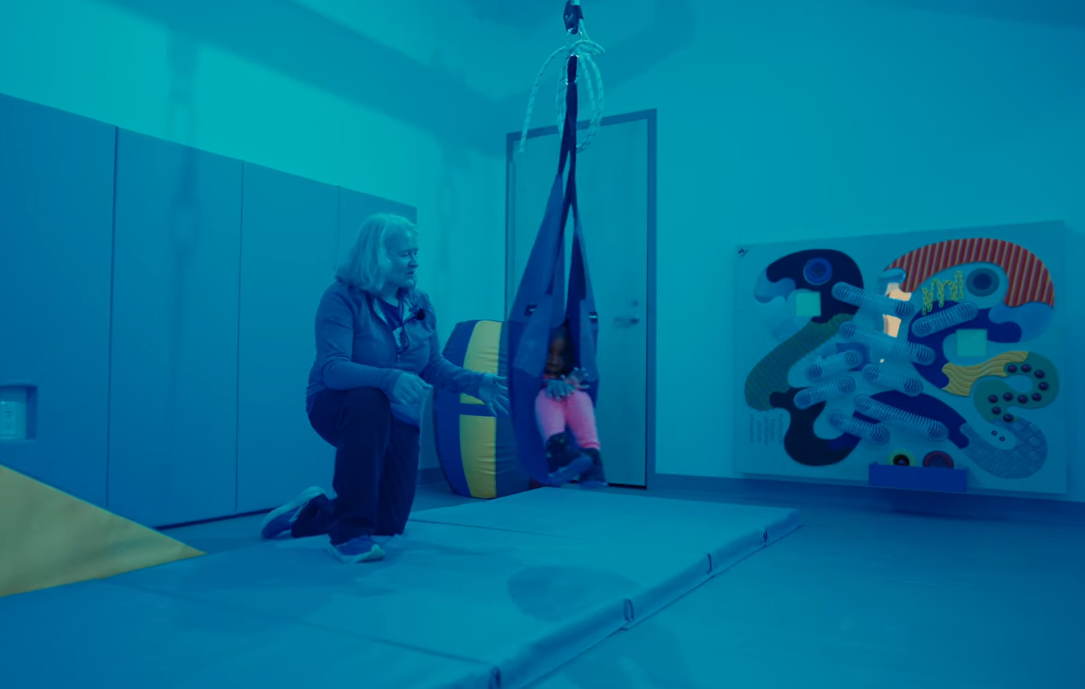 A child uses a suspended sensory swing with support from an educator in a sensory gross motor room designed to promote movement, regulation, and sensory exploration.