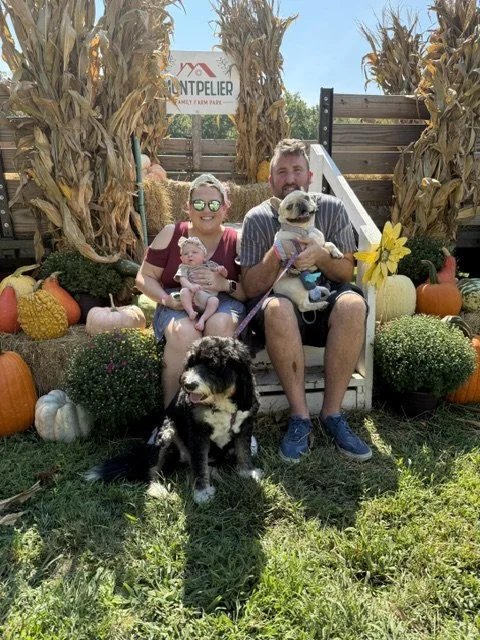 A family of four with two dogs sitting on a white chair outdoors at a pumpkin patch, surrounded by fall decorations and pumpkins.