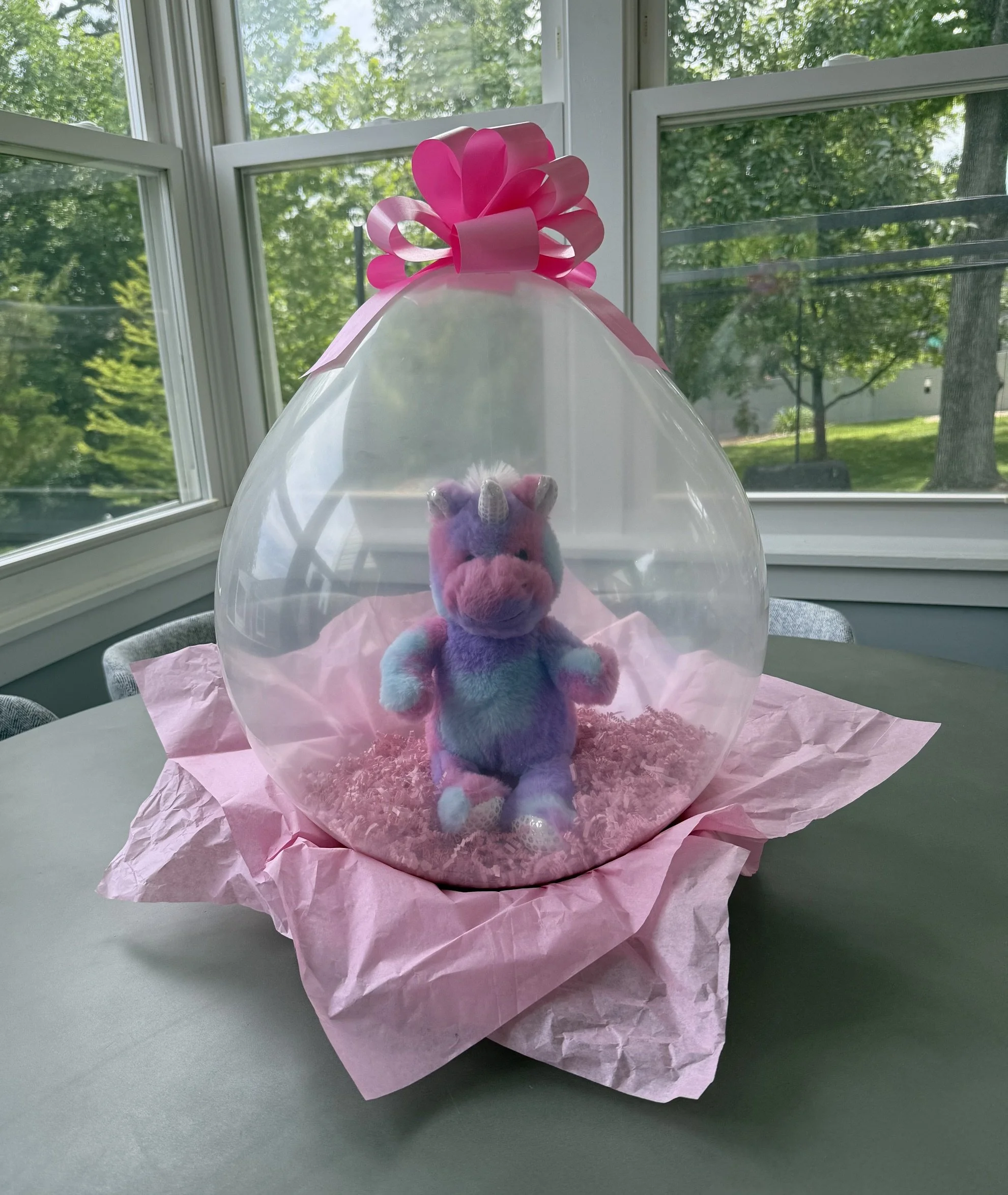 Colorful plush unicorn toy inside a clear plastic balloon, decorated with pink tissue paper and a pink bow, set on a table near windows with greenery outside.