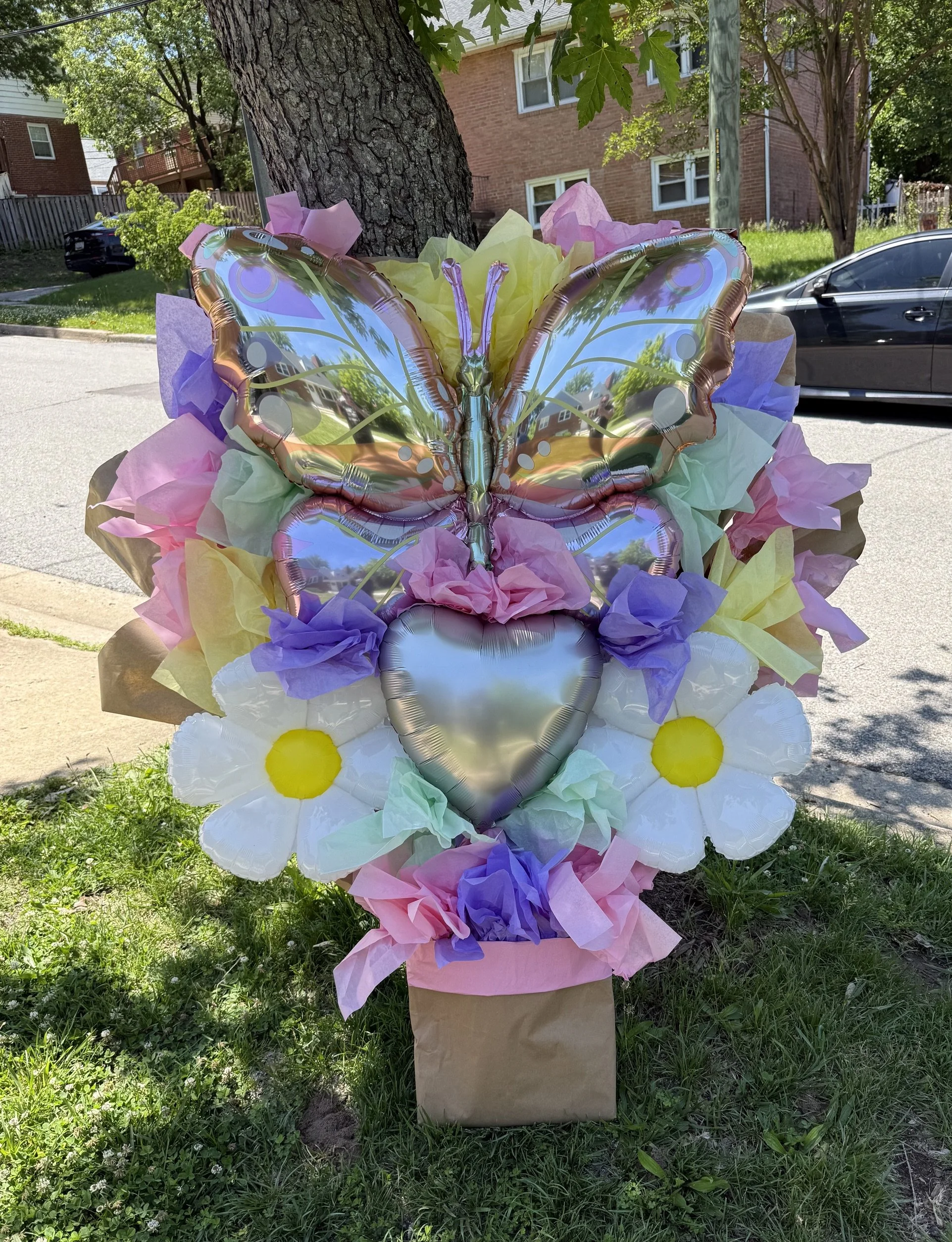 Colorful butterfly-shaped foil balloon surrounded by pastel-colored tissue paper and artificial flowers, attached to a yard sign with a brown paper base, set outdoors on a grassy area near a tree and residential building.