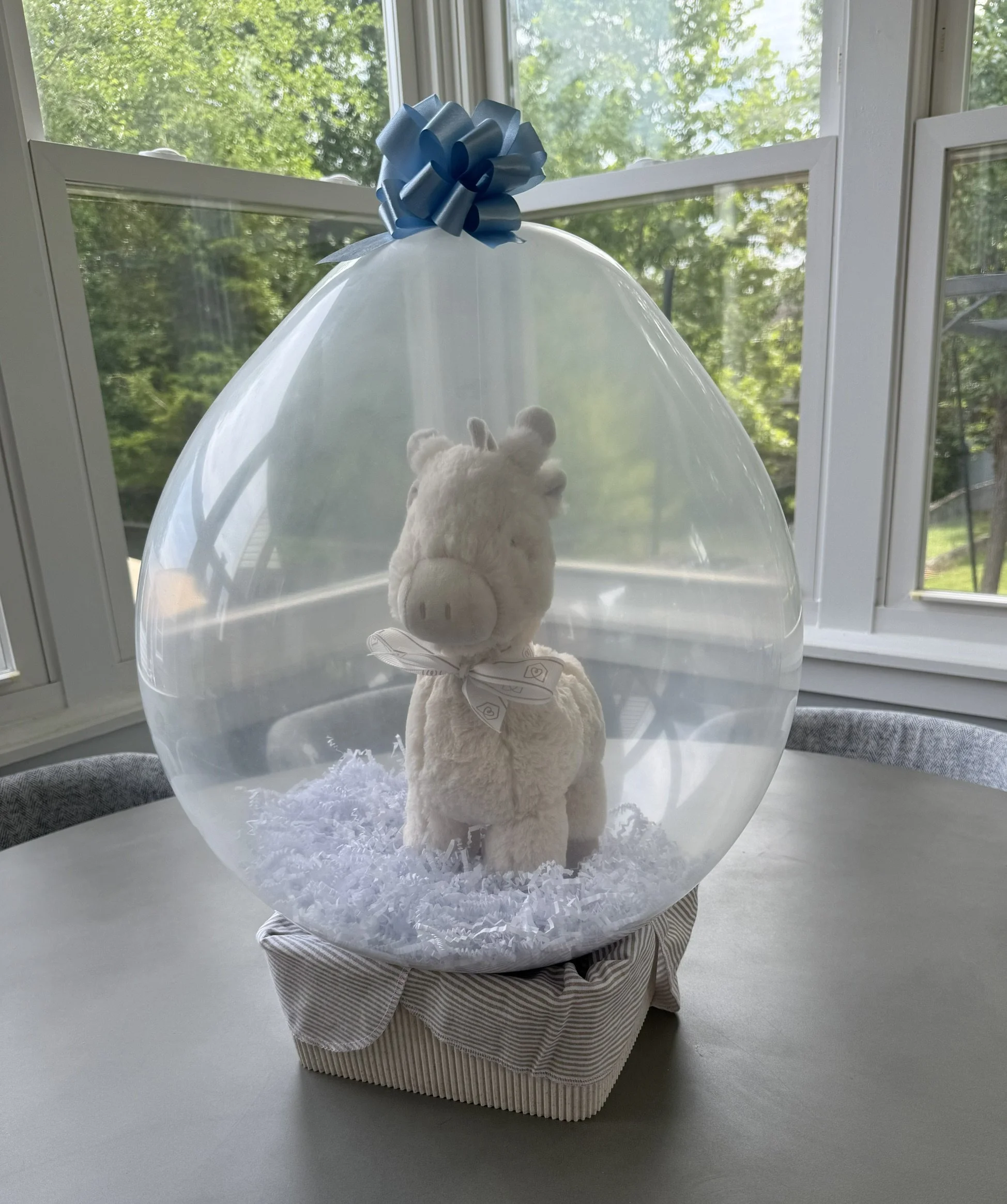 A plush stuffed animal bear wrapped in a white towel inside a clear balloon, with a blue ribbon bow on top, sitting on a round cushioned base, placed on a white windowsill with green trees outside.