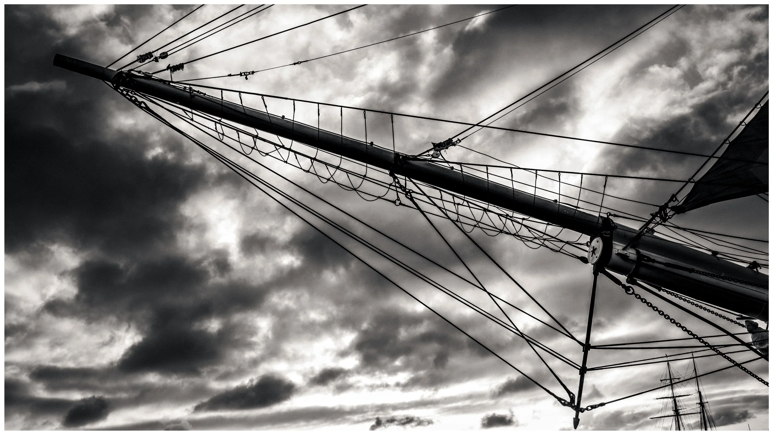 A black and white photo of a ship's mast with numerous riggings against a cloudy sky.