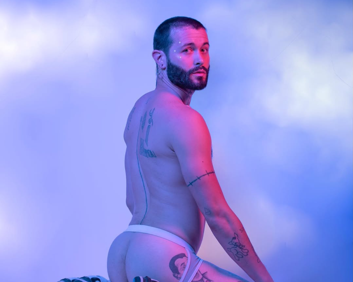A naked man with tattoos kneeling with his back to the camera, looking over his shoulder at the viewer against a cloudy sky background, illuminated by pink and blue lighting.