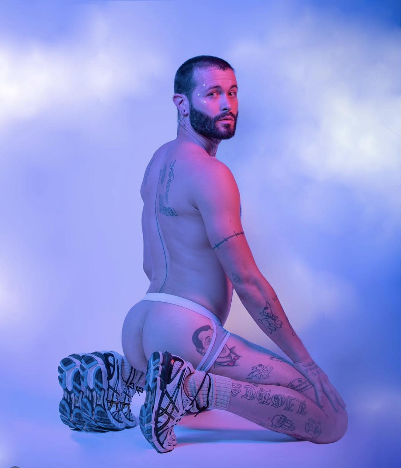 A shirtless man with tattoos kneels on the floor, wearing colorful sneakers and patterned socks, against a cloudy, colorful background with pink and blue hues.
