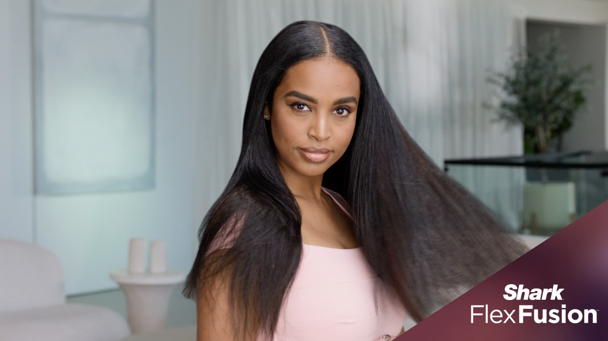 A woman with long, straight black hair and a light pink top in a modern, light-colored room with minimal decor and a potted plant in the background.