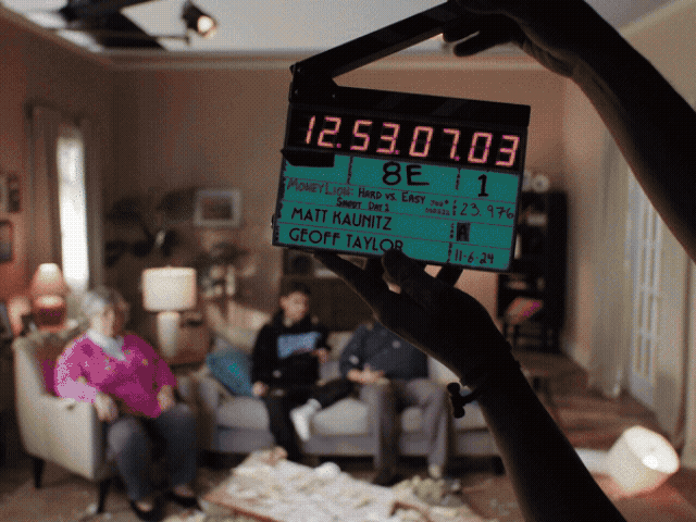 Film slate with timecode and scene details, held in front of a living room with three people sitting on a couch.